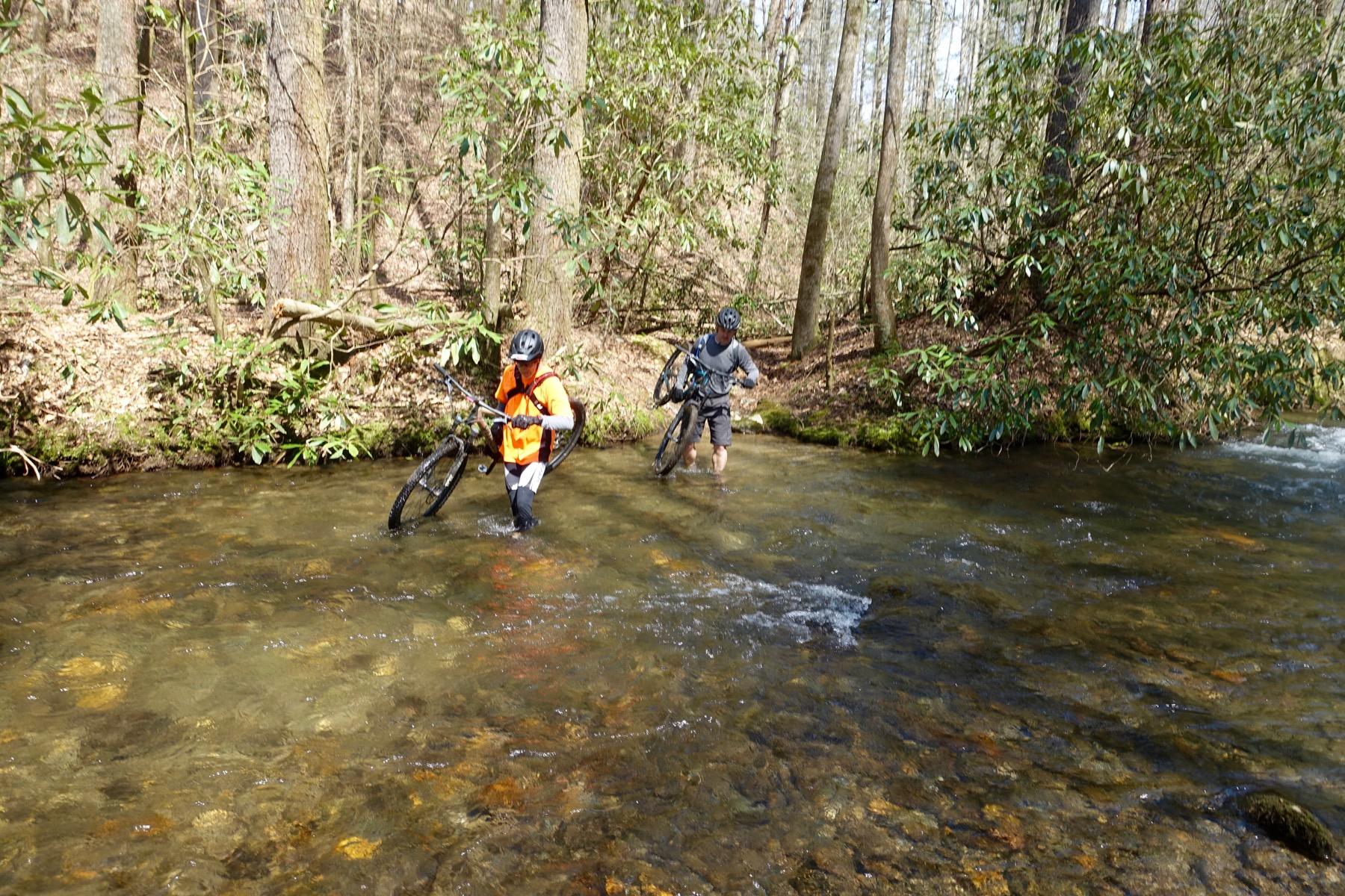 Two mountain bikers wade through a clear stream in a wooded area, carrying their bikes. One rider, wearing an orange shirt, is knee-deep in the water, while the other, dressed in a dark shirt and shorts, carefully steps through the flow. Surrounding them are tall trees and lush greenery, indicating a natural, outdoor setting. Pinhoti Trail: Mountaintown Creek Segment mountain bike trail.