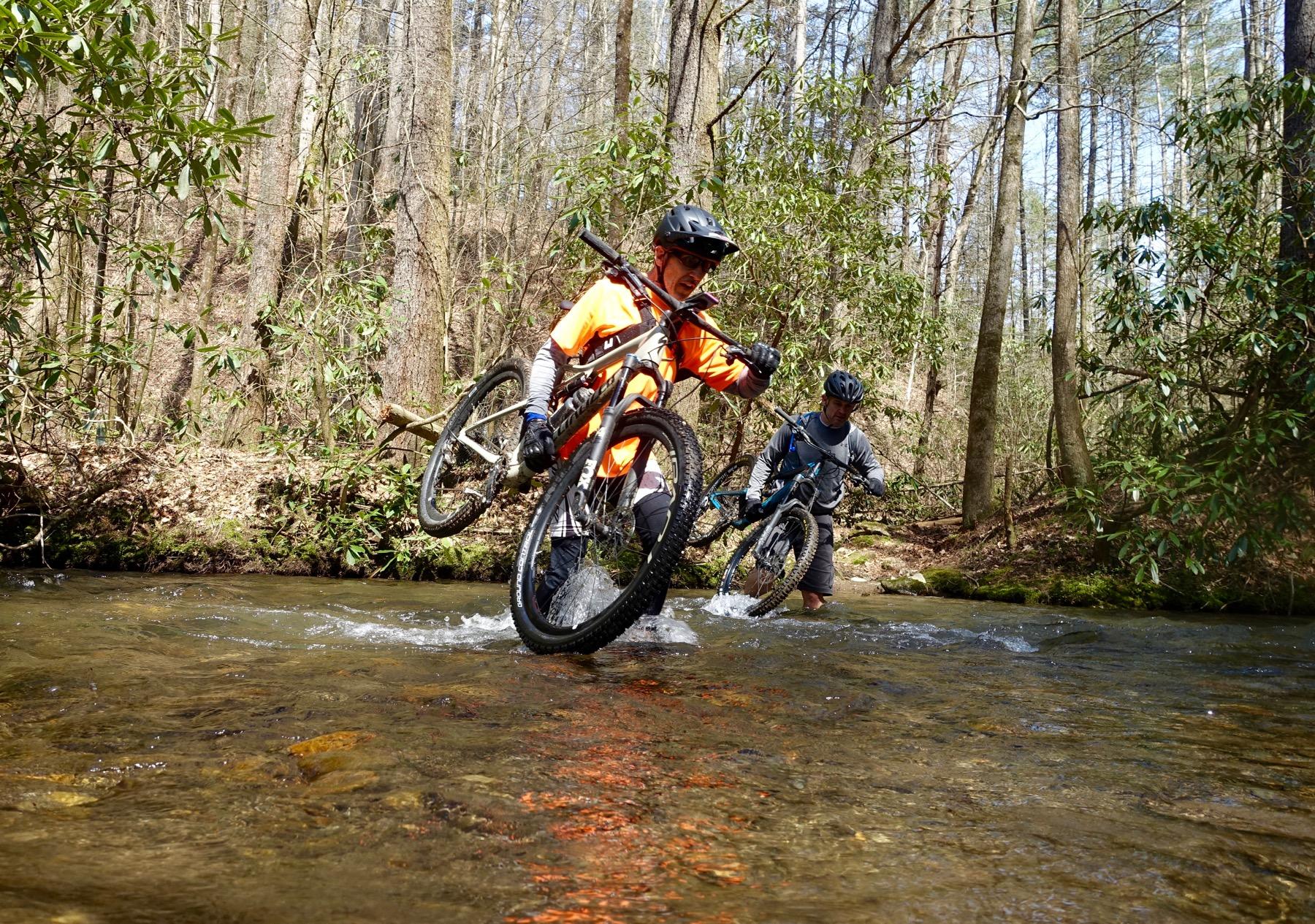 Two mountain bikers navigate a shallow stream, carrying their bikes. One rider, wearing an orange shirt and helmet, lifts his bike as he wades through the water, while the other rider, in a gray long-sleeve shirt, walks beside him. The scene is set in a wooded area with trees and greenery in the background, suggesting a sunny day. Pinhoti Trail: Mountaintown Creek Segment mountain bike trail.