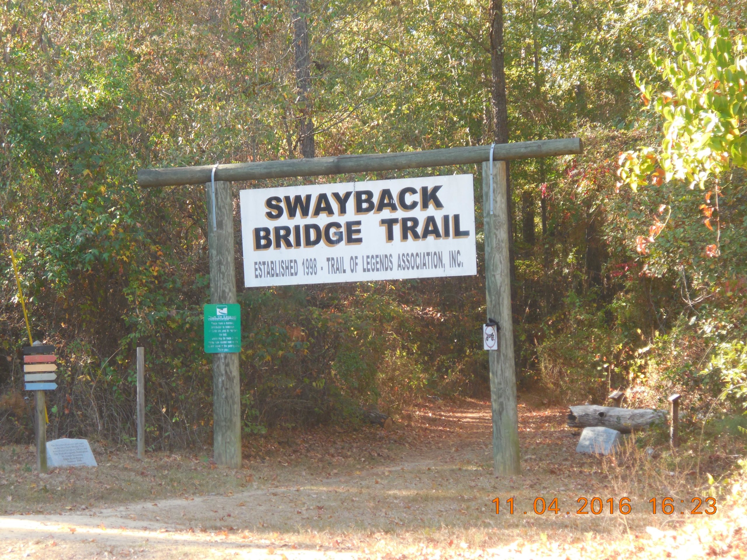Sign for Swayback Bridge Trail, established in 1998 by the Trail of Legends Association, surrounded by trees and greenery, with trail markers and a seating area nearby. Swayback Bridge Trail mountain bike trail.
