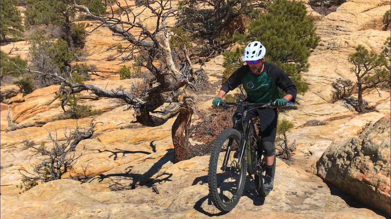 A mountain biker navigating rocky terrain, wearing a helmet and riding gear, in a natural outdoor setting with sparse trees and rugged landscape. Red Mesa mountain bike trail.