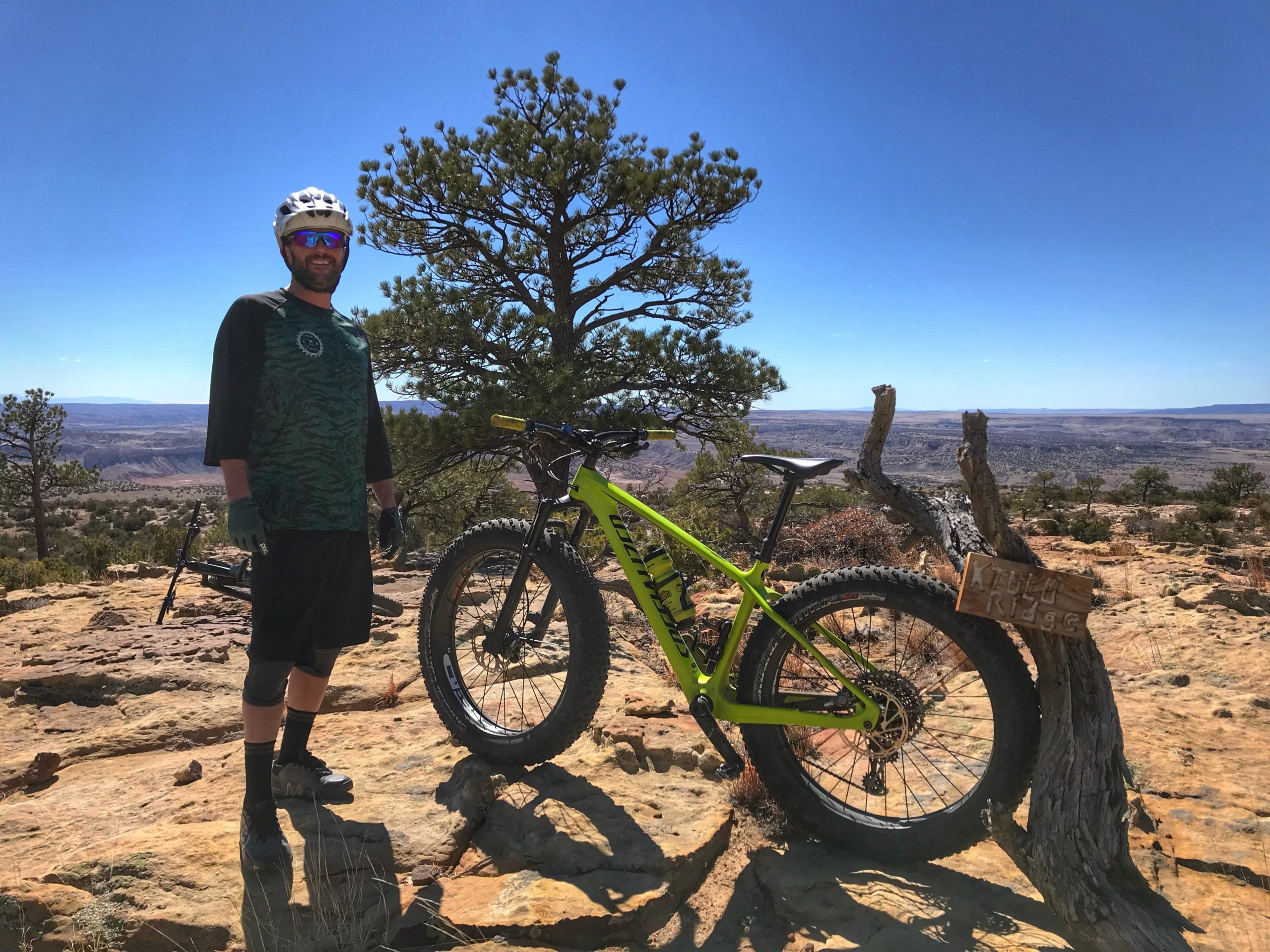 A mountain biker standing on a rocky outcrop with a vibrant green bike beside him. He is wearing a helmet, sunglasses, a black and green shirt, and shorts. In the background, there is a panoramic view of a desert landscape with distant mountains and a clear blue sky. A wooden sign labeled "Kith Ridge" is partially visible near the bike. Red Mesa mountain bike trail.
