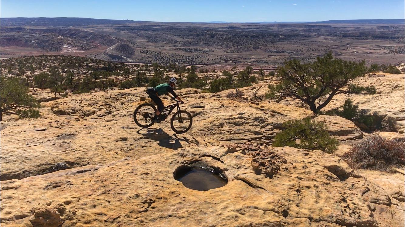 A mountain biker navigating rocky terrain on a sunlit plateau, with a vast landscape of valleys and hills in the background, dotted with trees. There is a noticeable rock formation featuring a circular indentation in the foreground. Red Mesa mountain bike trail.
