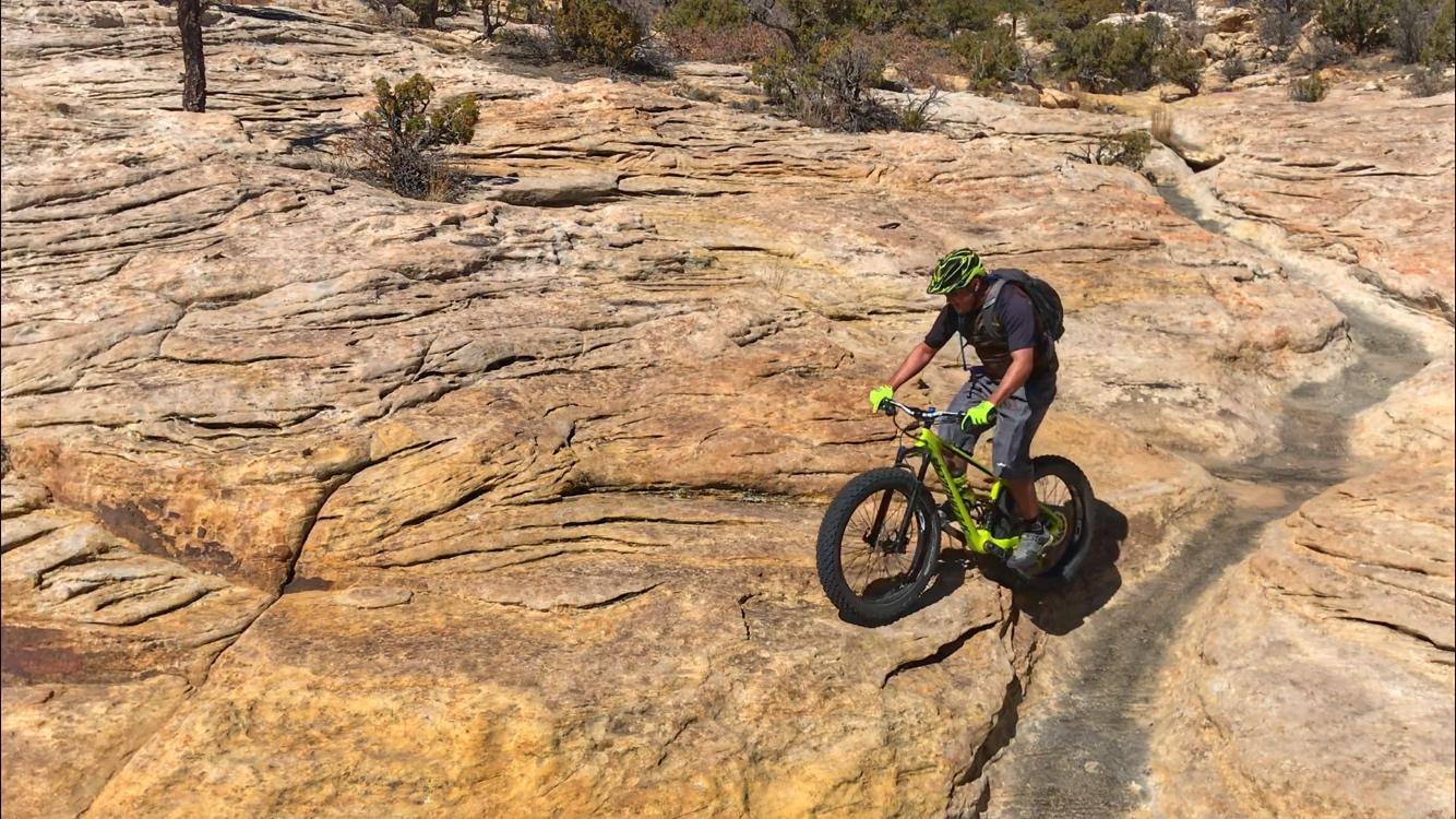 A mountain biker skillfully navigating rough, rocky terrain, riding a green fat bike on a sunlit trail surrounded by sparse vegetation and sandstone formations. Red Mesa mountain bike trail.
