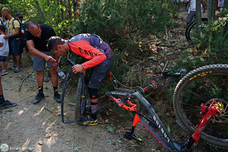 A mountain biker in an orange and gray jersey is crouched down next to their bike, inspecting a damaged tire. Another person is pointing at the bike while holding a water bottle. The scene is set in a wooded area with spectators in the background. The surrounding environment includes trees and a dirt path.