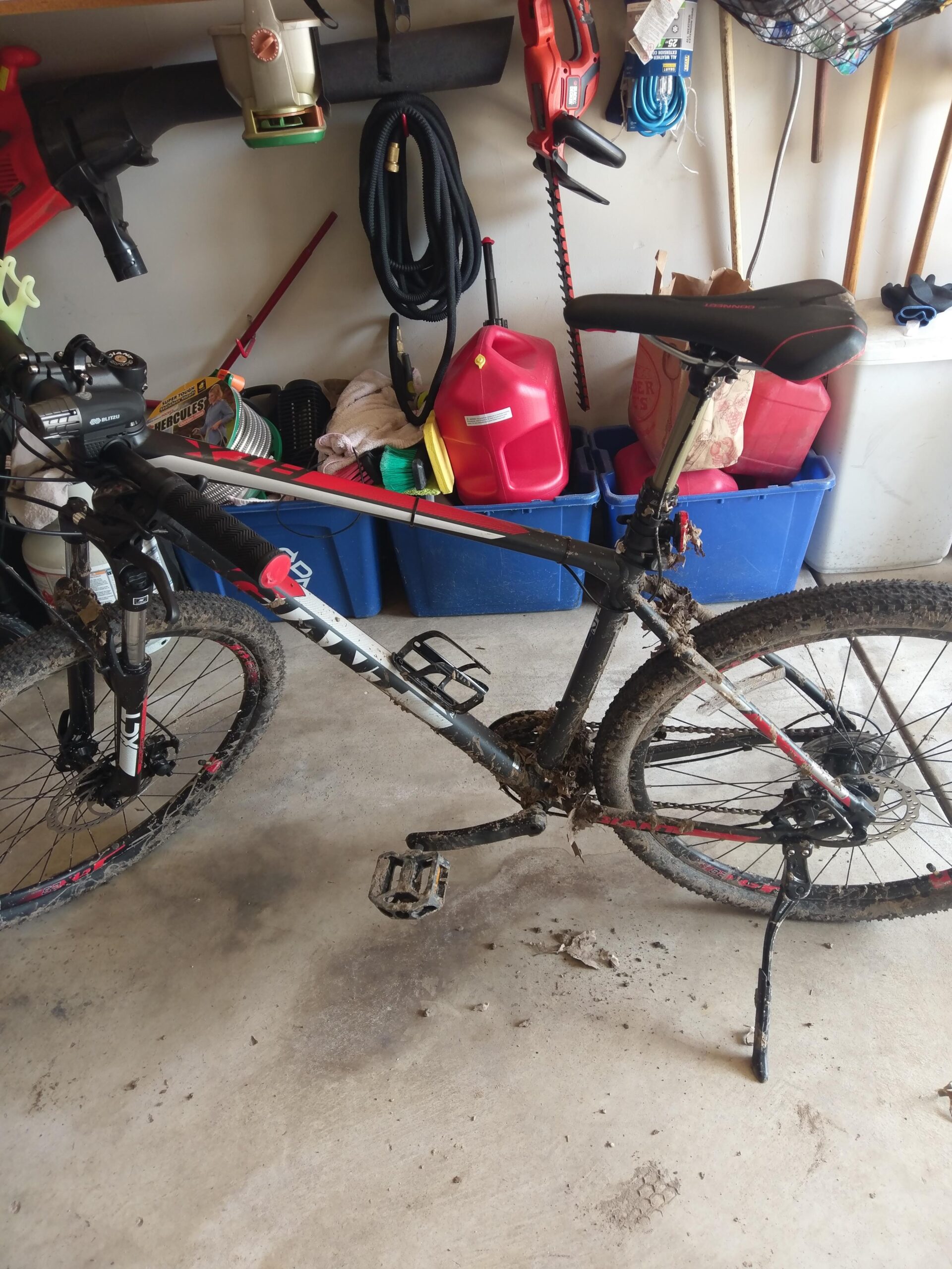 Giant ATX 27.5 2: A mountain bike with a muddy frame and wheels is parked in a garage. Various tools and equipment are visible in the background, including a leaf blower, hedge trimmer, and a gas canister, along with plastic storage bins. The garage floor shows muddy stains, indicating recent use.
