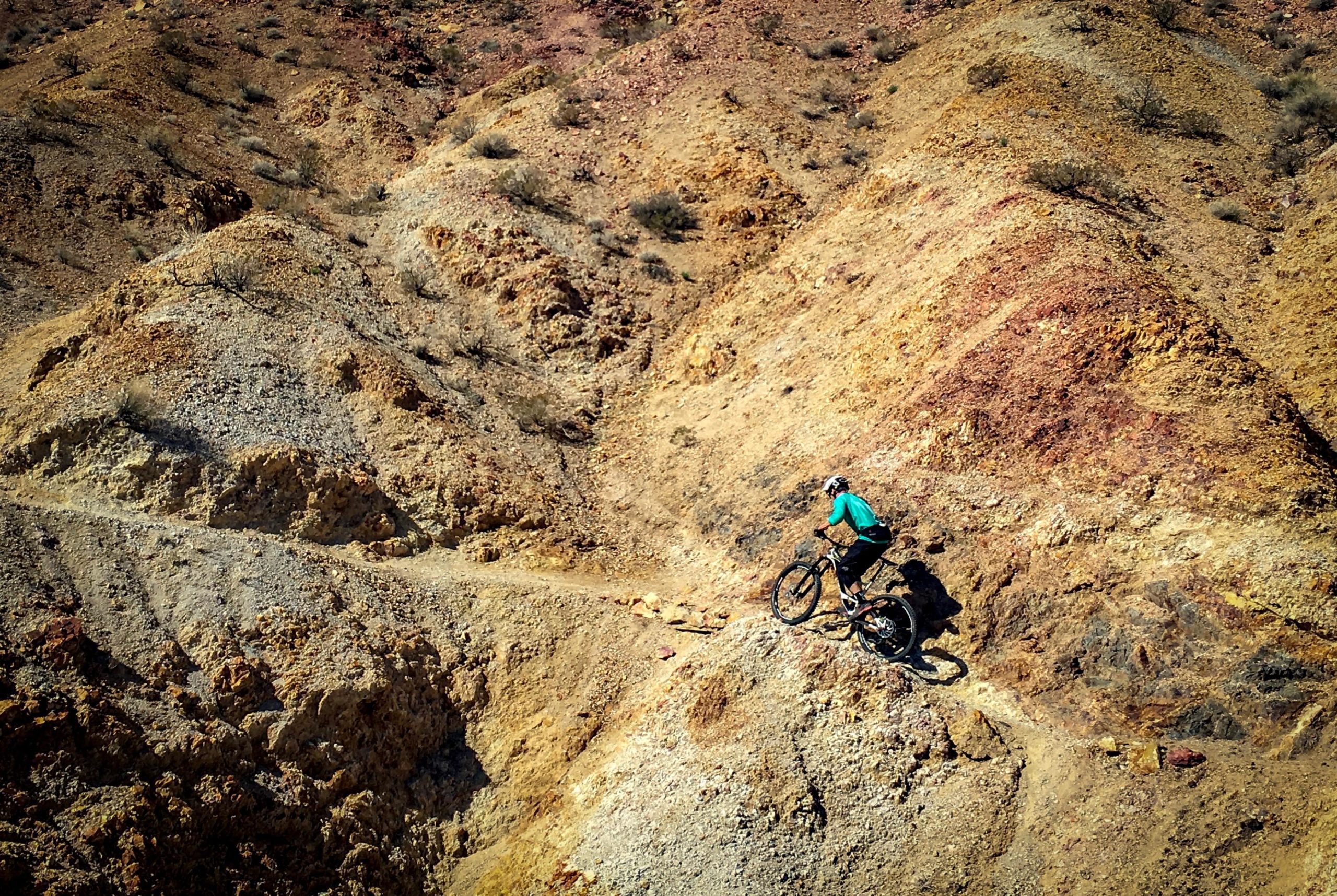 A mountain biker navigating a rocky, rugged terrain with slopes of red and yellow earth under a clear blue sky. The cyclist is wearing a teal shirt and black shorts, focused on maneuvering down the steep path. Sparse vegetation is visible amidst the rocky landscape. Bootleg Canyon mountain bike trail.