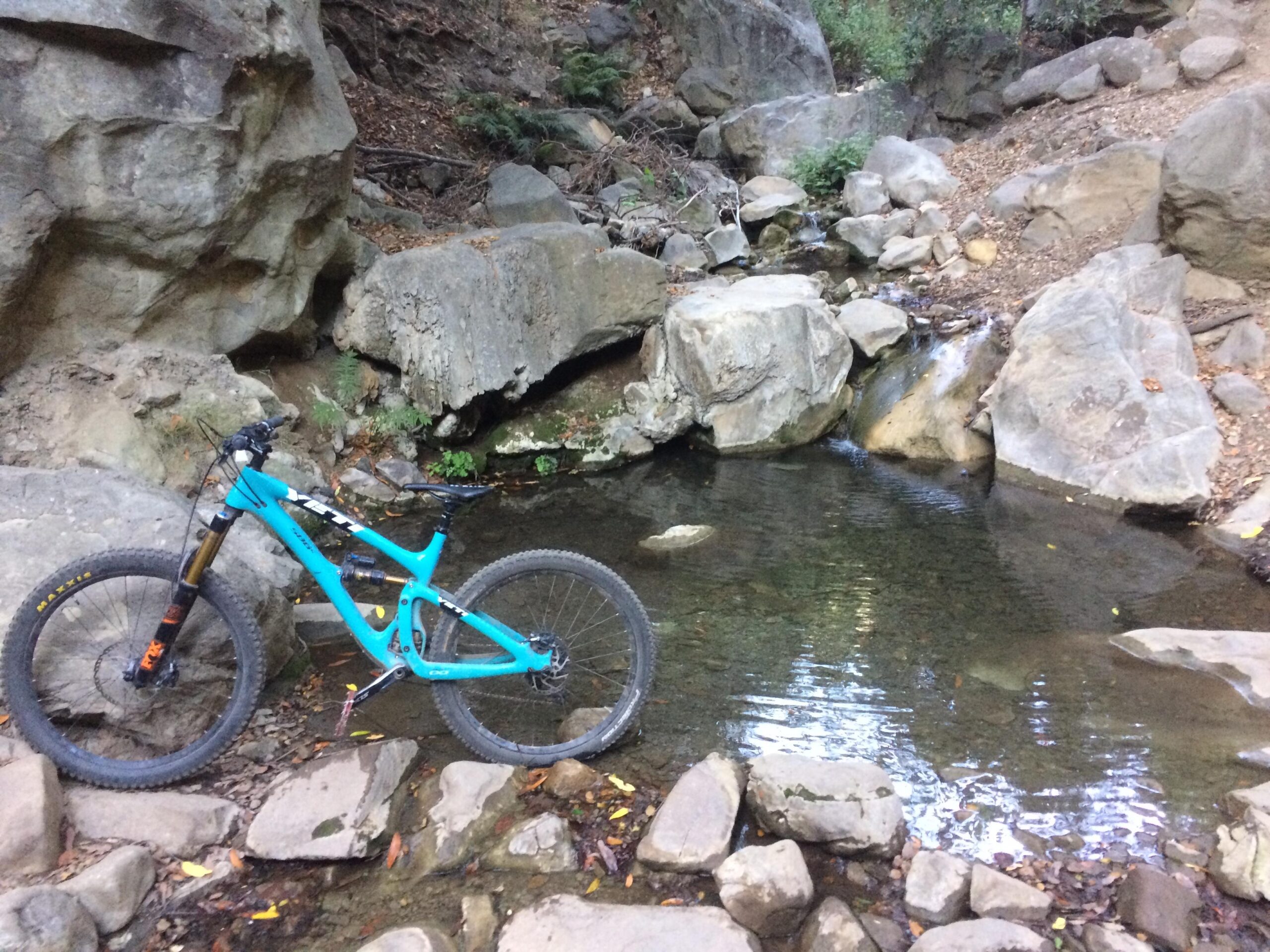 Yeti SB6: A bright blue mountain bike stands beside a tranquil stream, surrounded by large rocks and greenery. The water reflects the natural landscape, creating a serene outdoor setting.