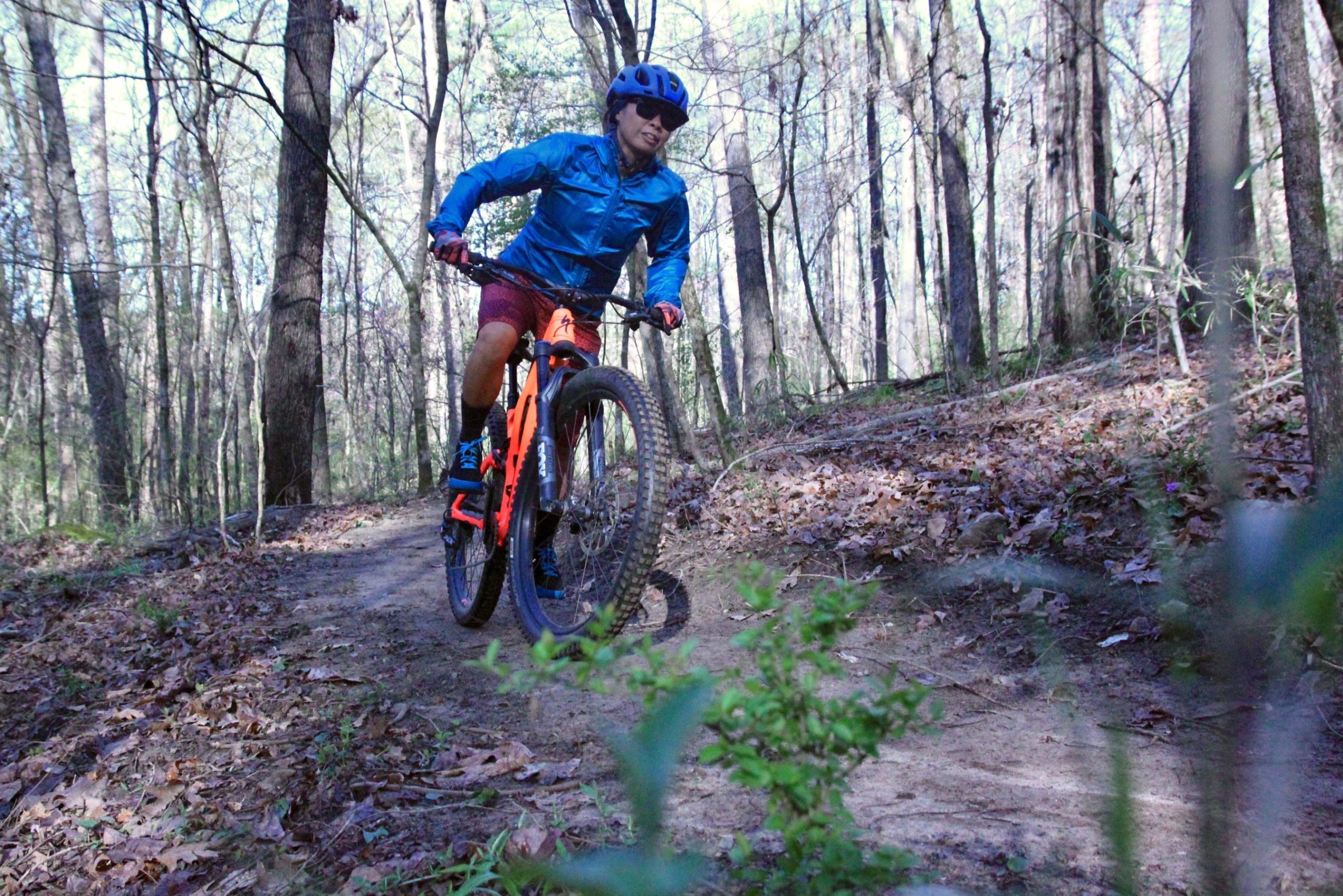 A person riding a mountain bike on a dirt trail surrounded by trees. The rider is wearing a blue jacket, helmet, and shorts, actively navigating the path with a slight lean forward. Sunlight filters through the trees, creating a dappled effect on the ground covered with fallen leaves. Southside Park mountain bike trail.