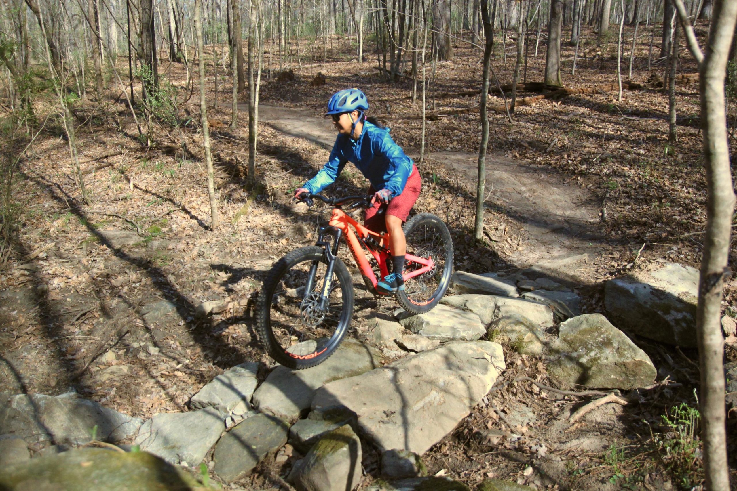 A young person wearing a blue jacket and red shorts rides a mountain bike over a rocky path in a wooded area. Sunlight filters through the trees, casting shadows on the ground covered with fallen leaves. Southside Park mountain bike trail.