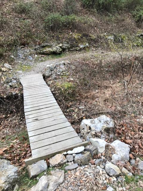 A wooden footbridge crossing a small dry creek bed, surrounded by rocky terrain and sparse vegetation. The landscape features fallen leaves and patches of grass, indicating a natural setting. Back 40 mountain bike trail.