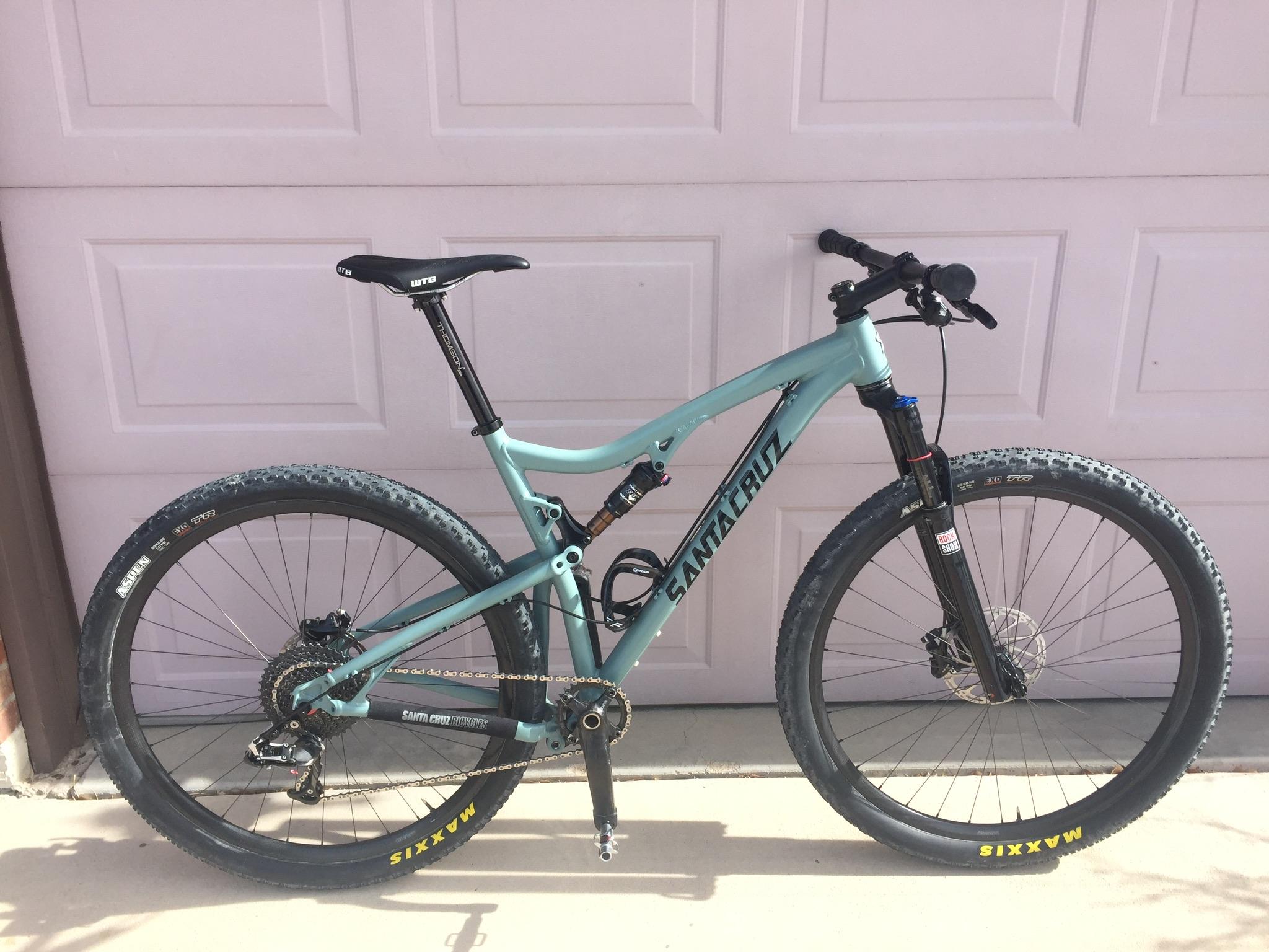 Santa Cruz Tallboy: A teal mountain bike leaning against a garage door, featuring wide tires and front suspension. The bike displays the brand name "Santa Cruz" prominently on the frame.