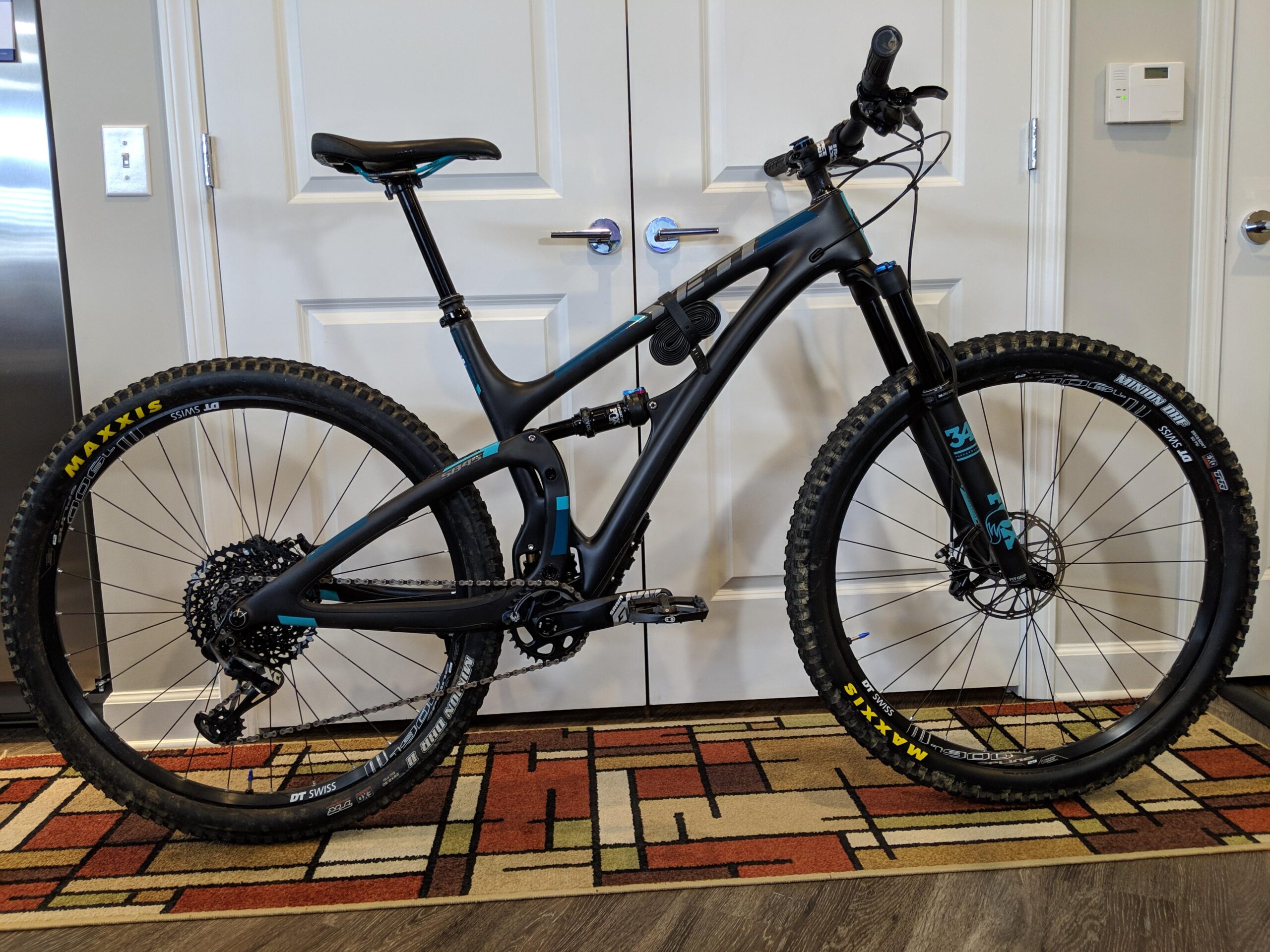 Yeti SB4.5c: A sleek black mountain bike with blue accents, featuring wide tires with Maxxis branding, positioned on a patterned rug. The bike has a modern frame design, front suspension, and gear components visible. It stands next to a door in a well-lit indoor space.