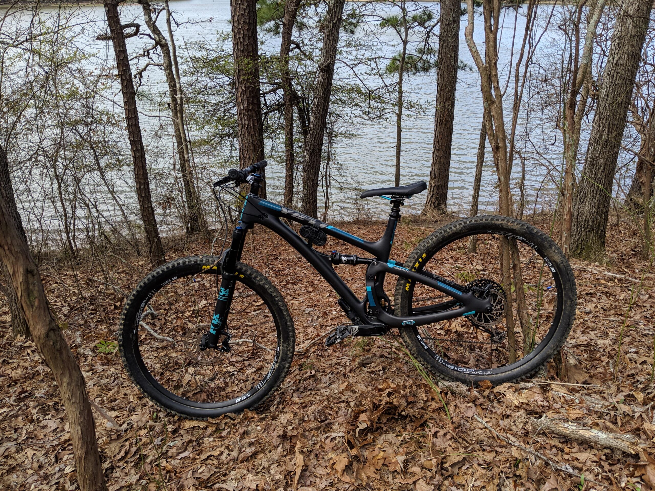 Yeti SB4.5c: A mountain bike parked on a leaf-covered trail beside a lake, surrounded by trees and shrubs. The bicycle features a black frame with blue accents and has thick tires suitable for off-road terrain.
