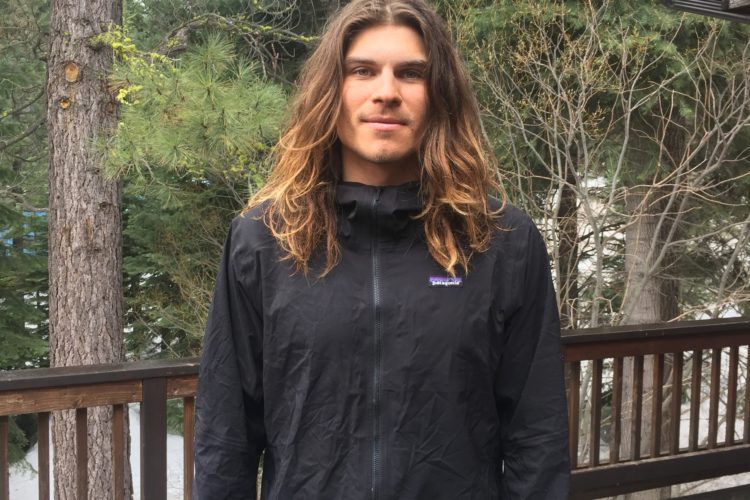 A person standing outdoors on a wooden deck, surrounded by evergreen trees. They are wearing a black jacket and have long, wavy hair. Snow is visible on the ground nearby, indicating a cooler climate. The setting appears to be in a natural, serene environment.