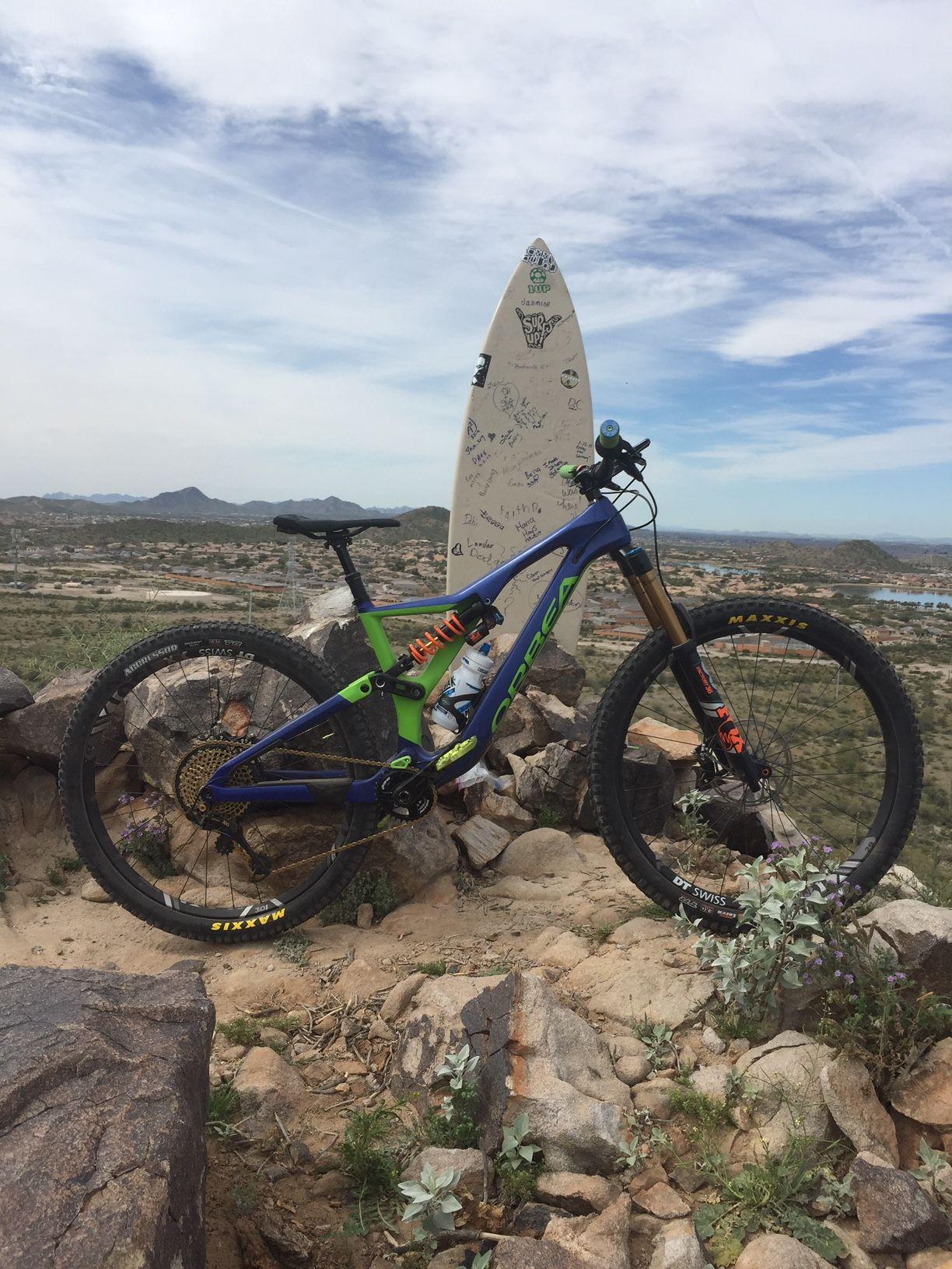 Orbea Rallon: A mountain bike positioned on rocky terrain with a surfboard in the background, featuring various stickers and graffiti. The scene captures a clear sky with wispy clouds and distant mountains, showcasing a blend of biking and surfing culture.