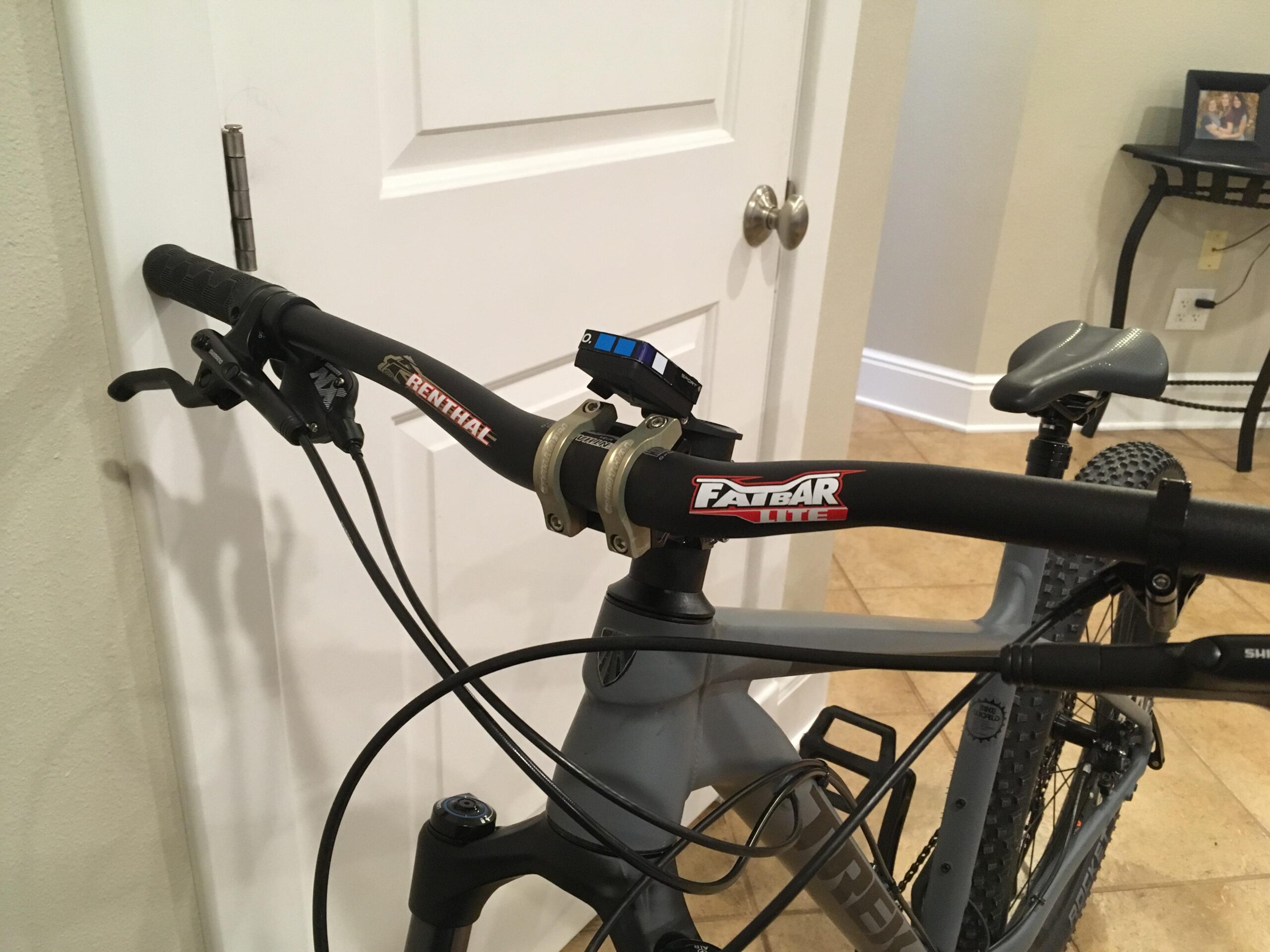 Trek Roscoe 8: Close-up of a bicycle handlebar featuring a Renthal sticker, a Fatbar Lite component, and a digital display. Cable components and brake levers are also visible. The background includes a door and a small table with a framed photo. The floor is tiled.