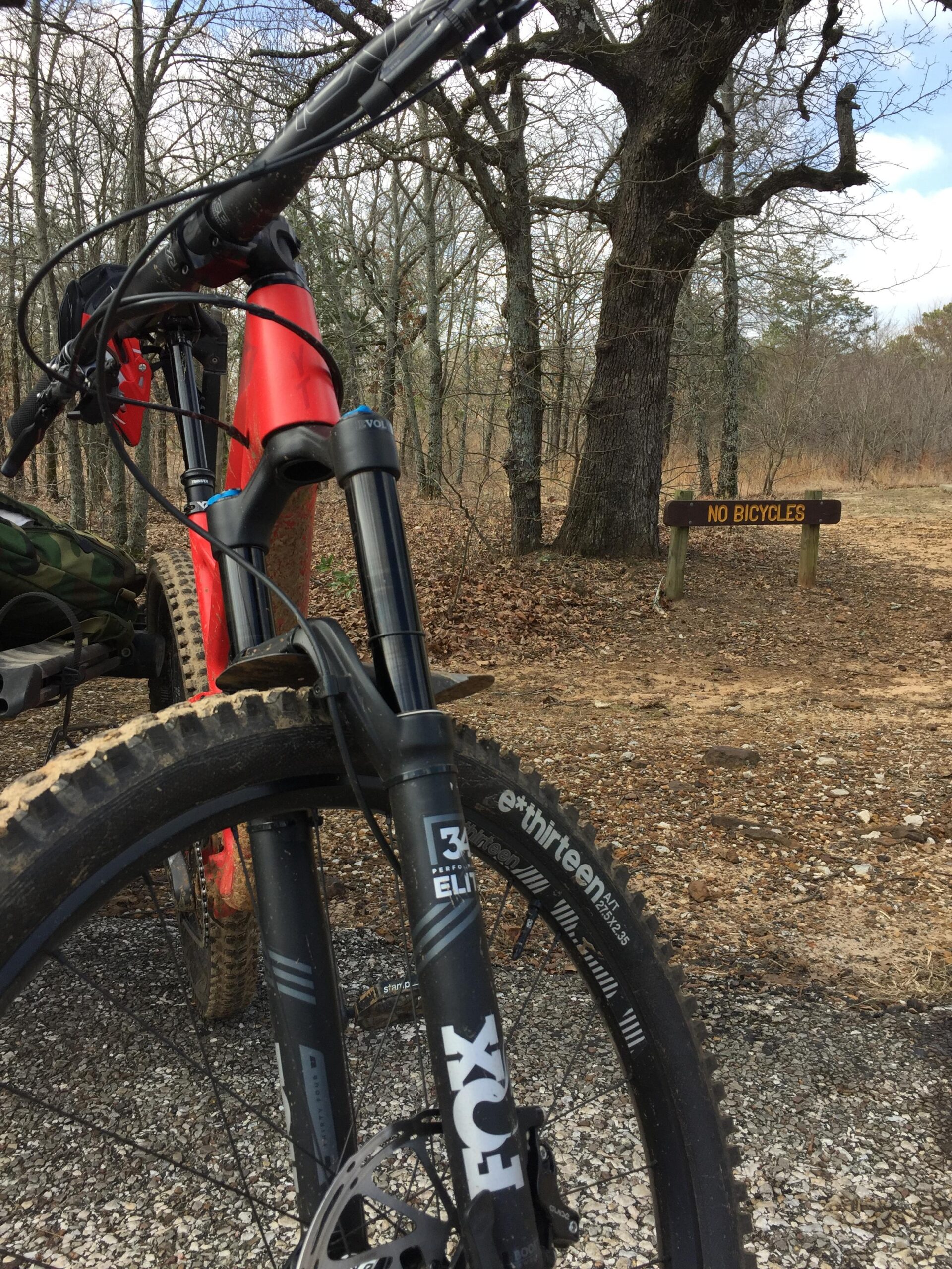 YT Industries CF Pro: A close-up view of a red mountain bike positioned on a gravel path, with a focus on the front wheel and fork. In the background, a wooden sign reads "No Bicycles," surrounded by bare trees and brown foliage, indicating the location is in a wooded area.