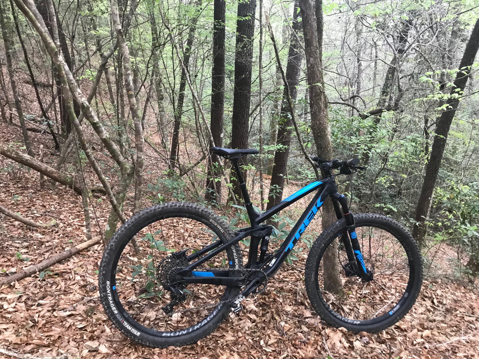 A mountain bike with blue accents is parked on a trail surrounded by trees and underbrush in a forested area. The ground is covered with fallen leaves, and the scene conveys a sense of nature and outdoor adventure. Bogue Chitto State Park mountain bike trail.