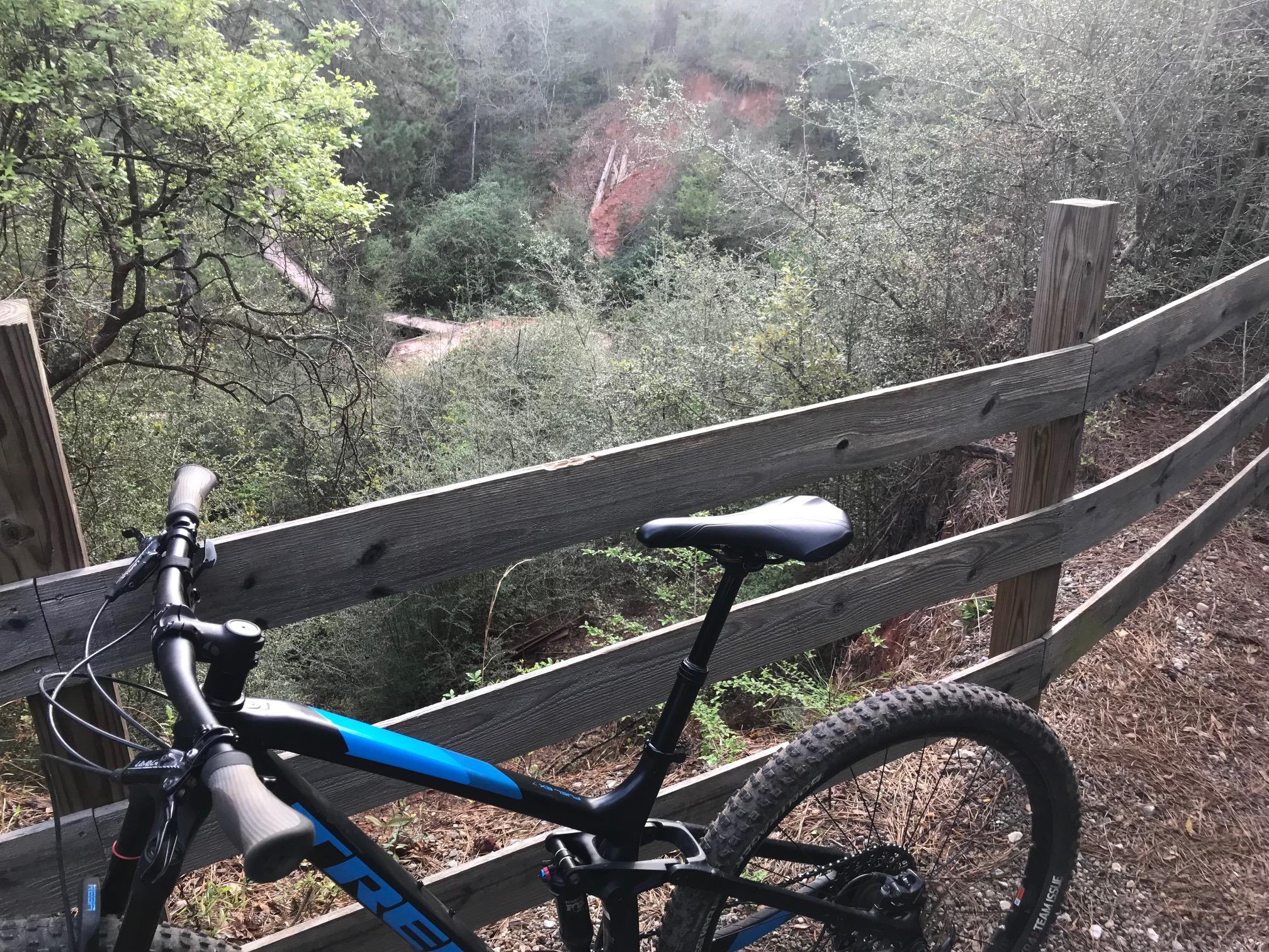 A mountain bike placed against a wooden fence, overlooking a lush, green landscape with a winding trail and scattered trees. The scene captures a serene outdoor moment in nature, ideal for cycling enthusiasts. Bogue Chitto State Park mountain bike trail.