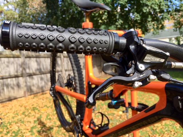Rocky Mountain Instinct C70: Close-up view of a bicycle handlebar featuring a textured black grip and a silver brake lever, with an orange frame partially visible in the background. The image captures autumn foliage on the ground, creating a seasonal atmosphere.