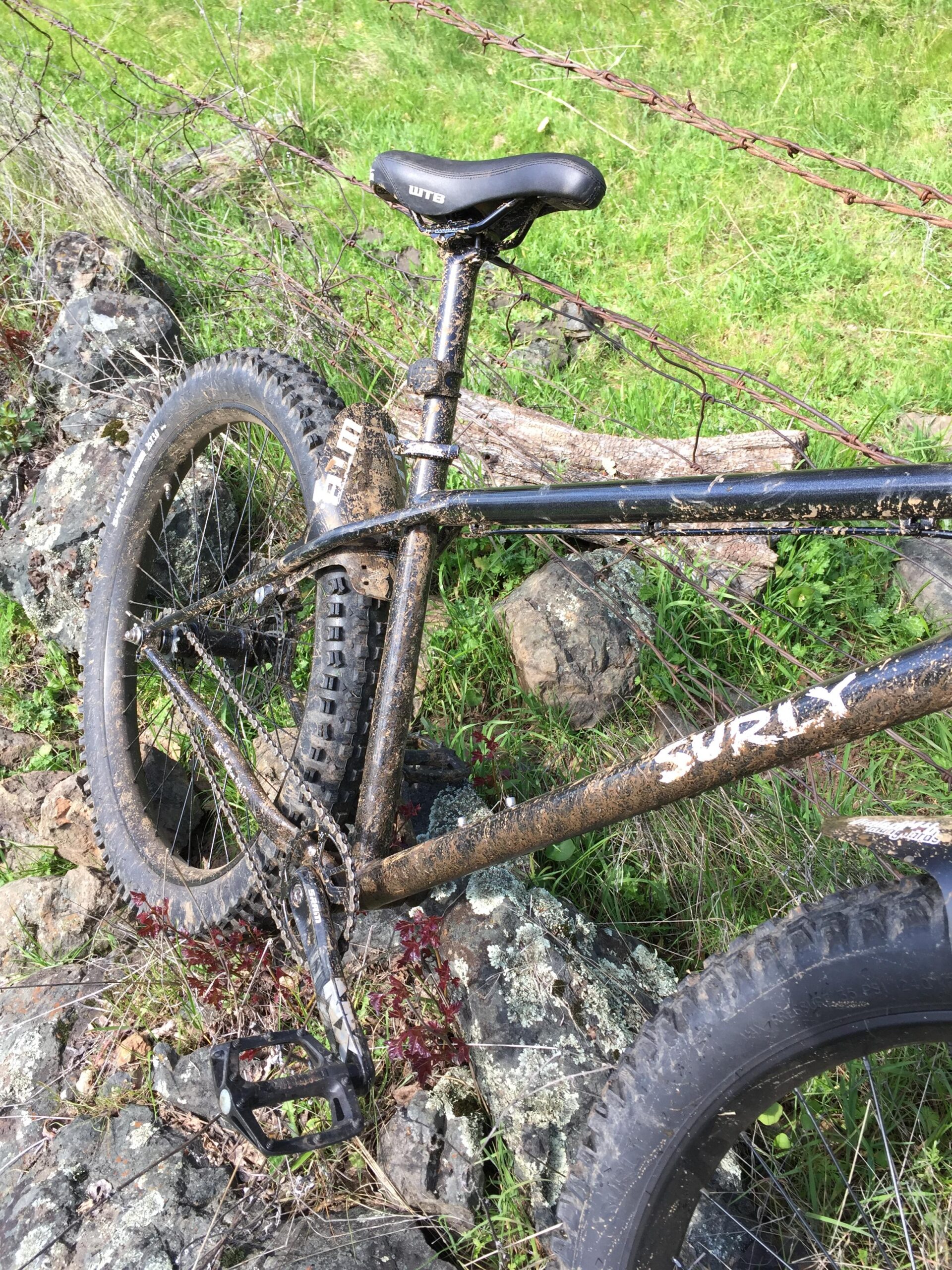 Surly Lowside: A close-up view of a muddy mountain bike resting on rocky terrain, partially leaning against a barbed wire fence. The bike features a black frame with a visible "Surly" logo, a black saddle, and knobby tires. The surrounding area is green with patches of grass and small plants.