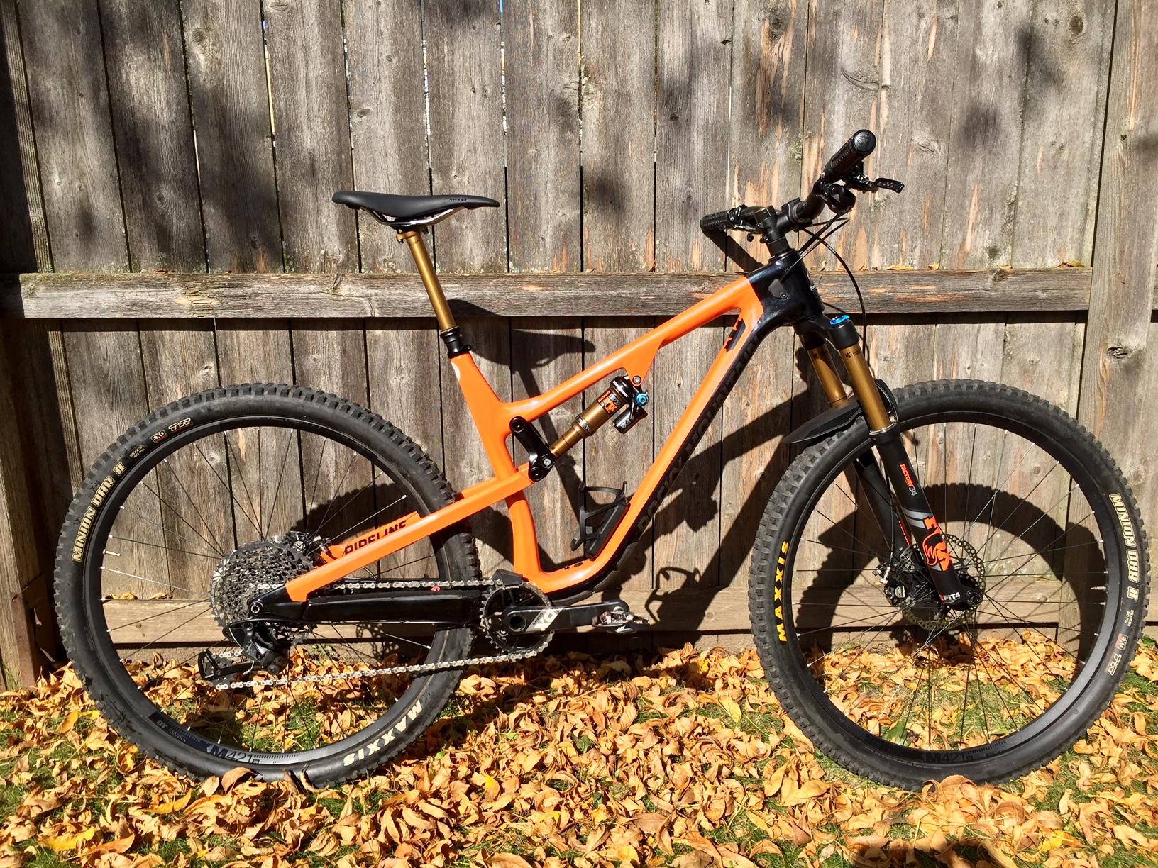 Rocky Mountain Instinct C70: A bright orange mountain bike leaning against a wooden fence, surrounded by fallen leaves. The bike features wide tires, a sturdy frame, and front suspension, showcasing its design for off-road biking.