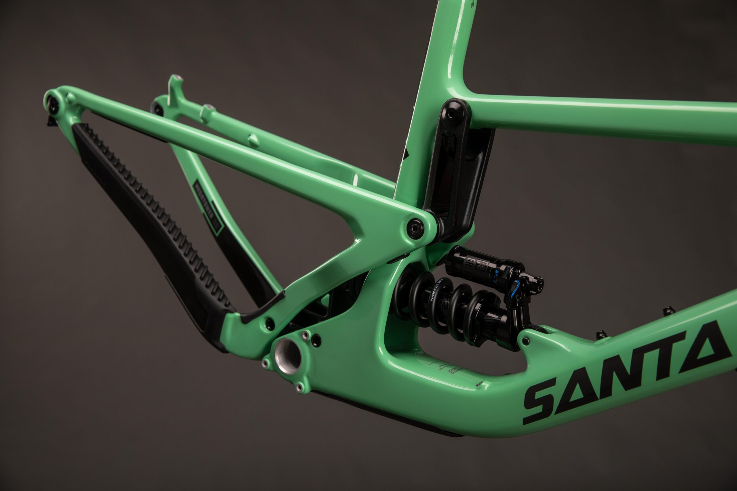 Santa Cruz Megatower: Image of a green mountain bike frame showcasing a suspension system, with a close-up view highlighting the shock absorber and structural design. The frame has a sleek, modern aesthetic with the brand name "SANTA" prominently displayed. The background is a dark shade, emphasizing the frame's color and details.