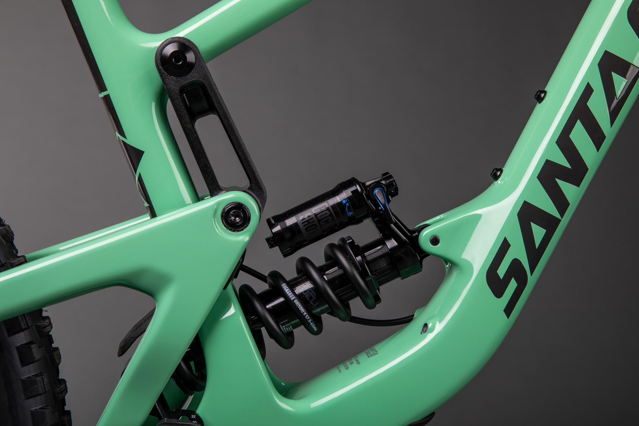 Santa Cruz Megatower: Close-up view of a mint green mountain bike frame featuring a black shock absorber. The image highlights the bike's suspension system, with visible components such as the coil spring and linkage. The background is a simple gray, emphasizing the bike's sleek design and color.