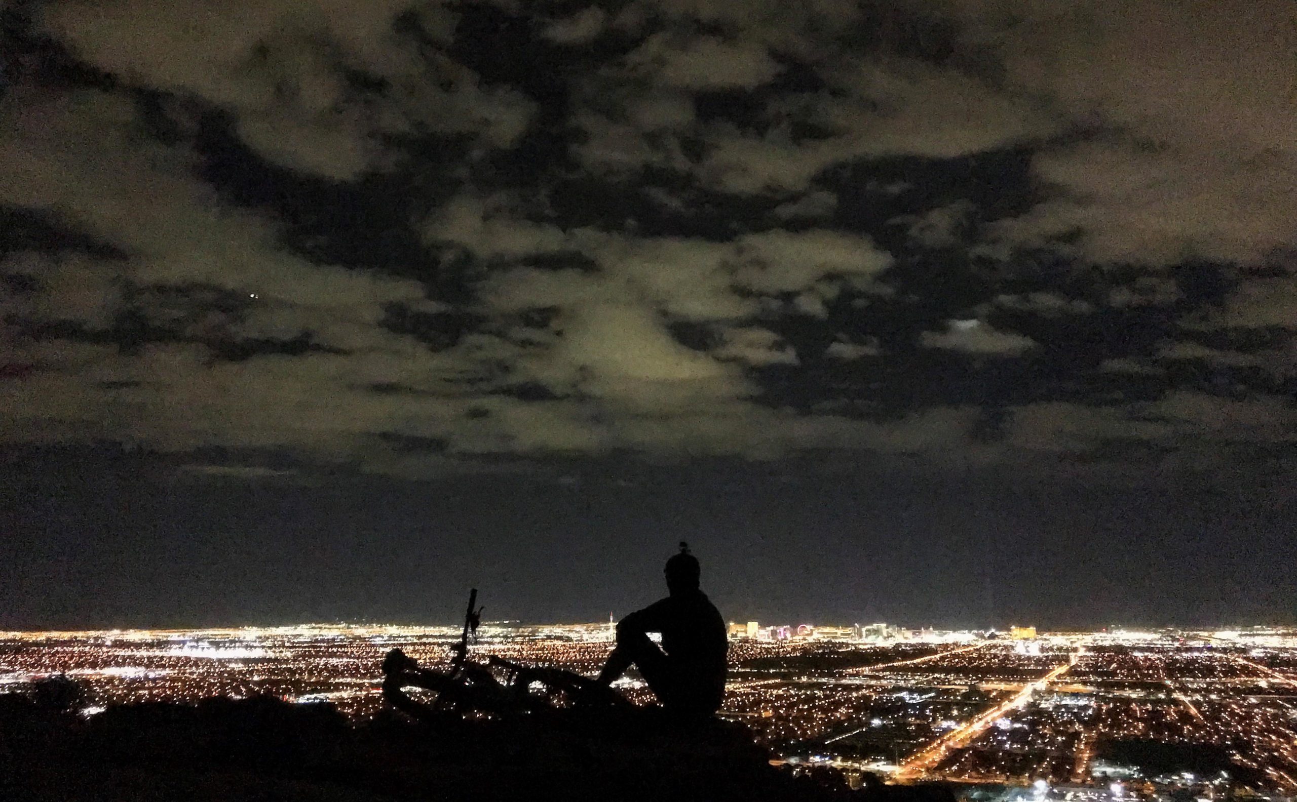 A person silhouetted against a night sky, sitting on a rocky ledge overlooking a city lit up with streetlights and buildings. Clouds drift across the sky, and the vibrant lights of the city create a striking contrast against the darkness. Bears Best mountain bike trail.