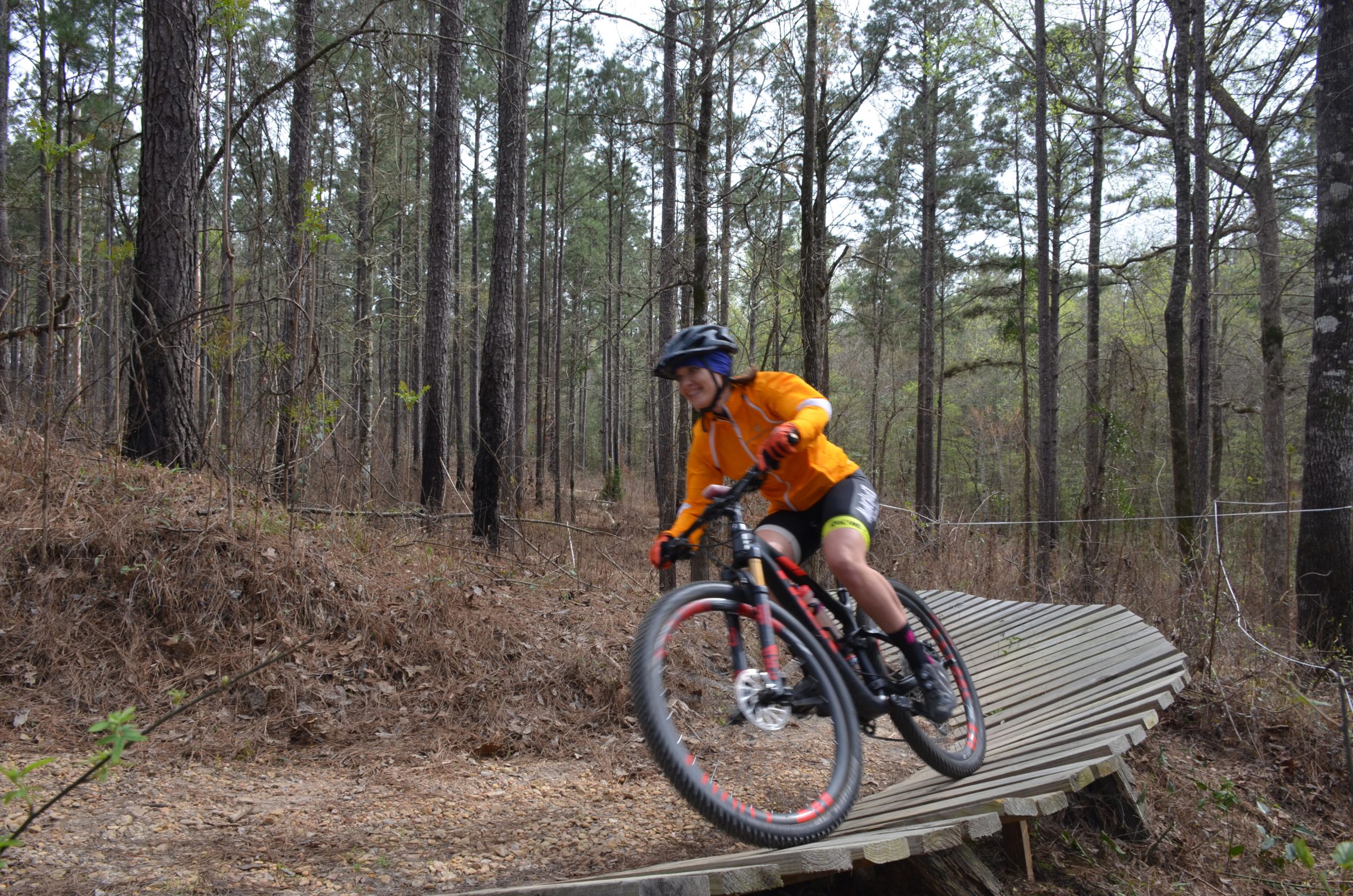 A person in an orange jersey and black shorts rides a mountain bike on a wooden track through a forested area. They are leaning into a turn, showcasing motion and action, with tall pine trees in the background and a trail of brown foliage beneath. Mt. Zion Bike Trails mountain bike trail.