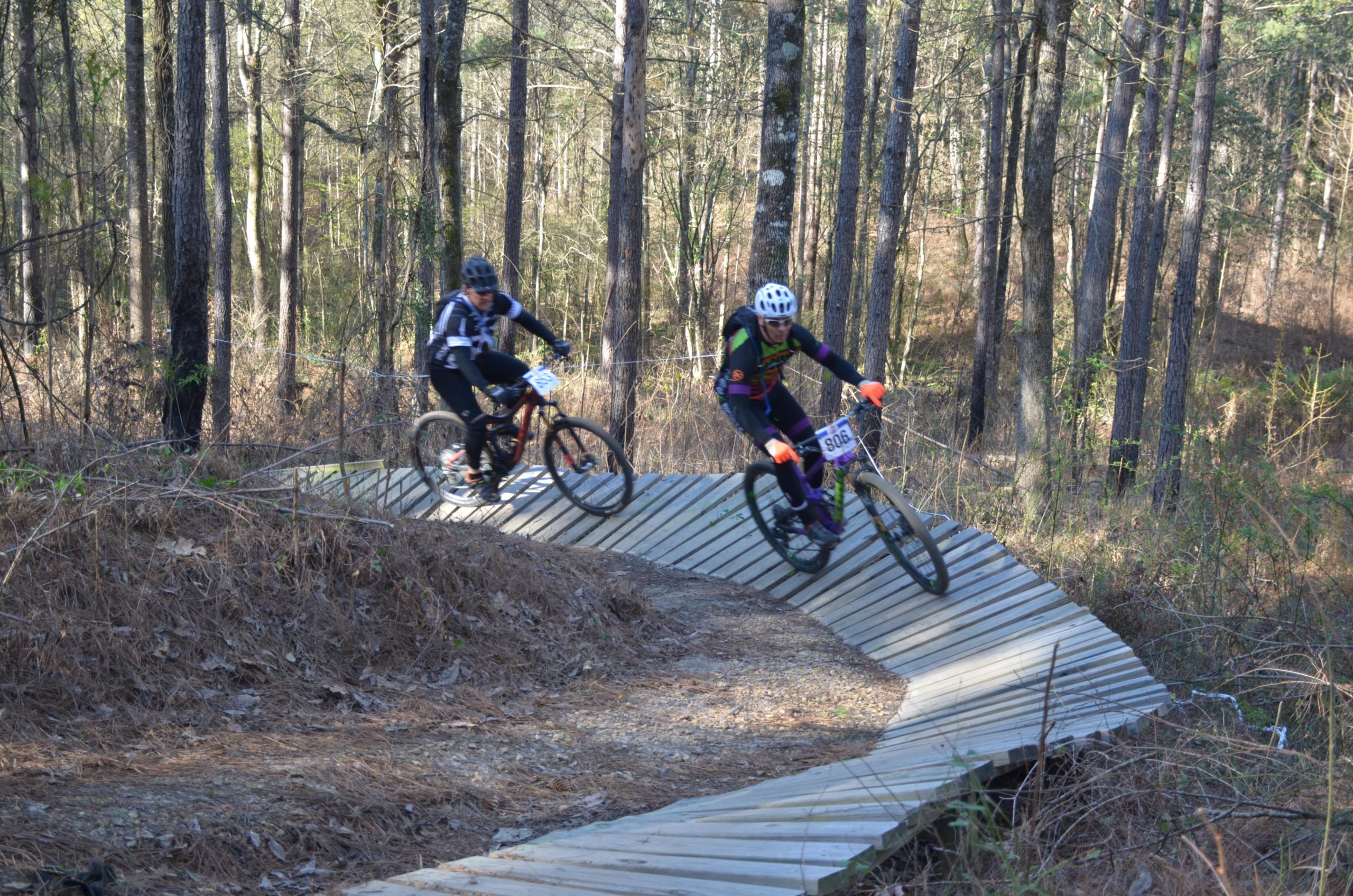 Two mountain bikers navigating a wooden trail in a forested area. One rider, wearing a black and white jersey, is positioned to the left, while the other, dressed in a colorful outfit with a helmet, is on the right. The background features tall trees and underbrush typical of a natural wooded environment. Mt. Zion Bike Trails mountain bike trail.