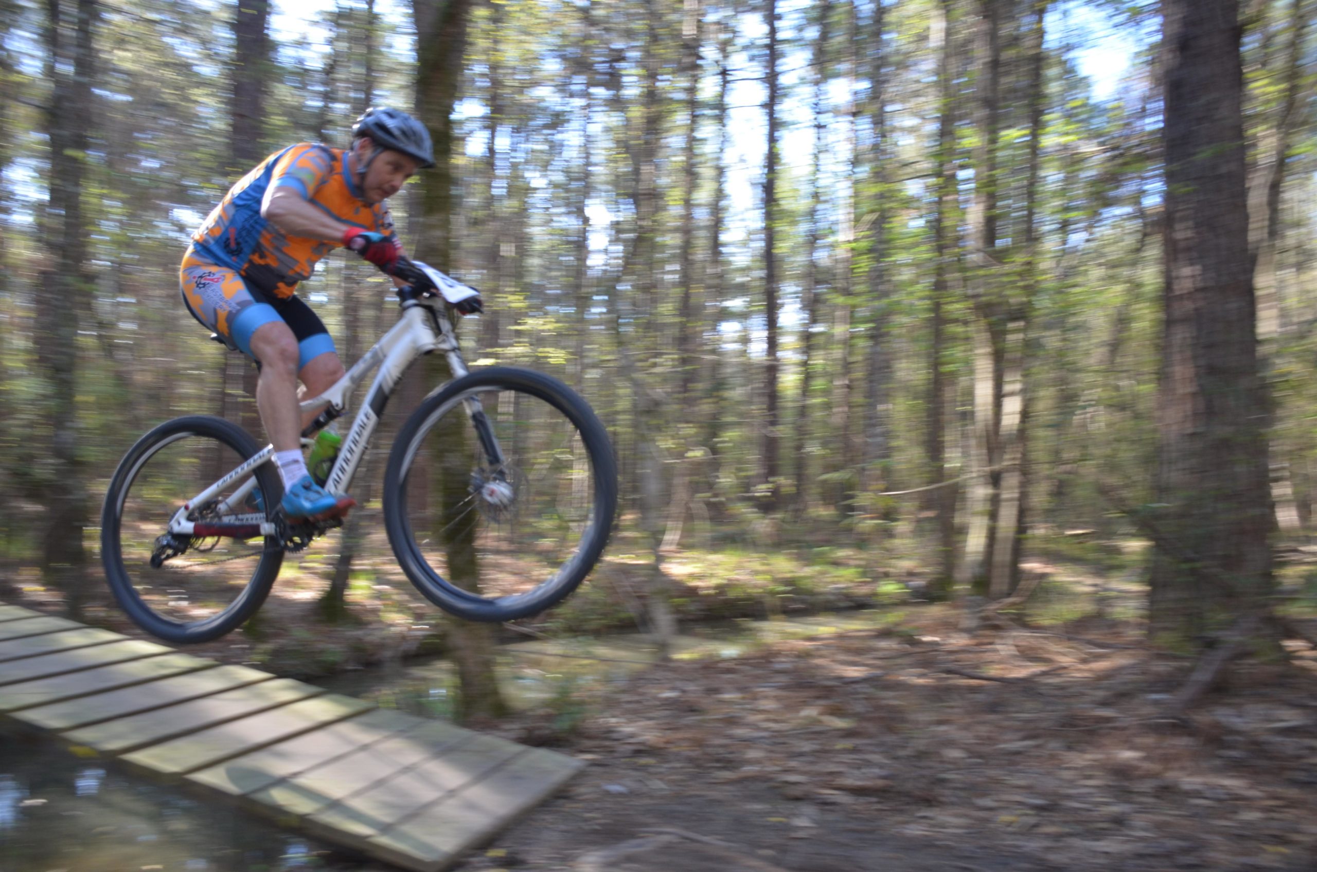 A mountain biker in colorful attire jumps off a wooden bridge while riding through a lush forest, with tall trees and dappled sunlight visible in the background. Mt. Zion Bike Trails mountain bike trail.