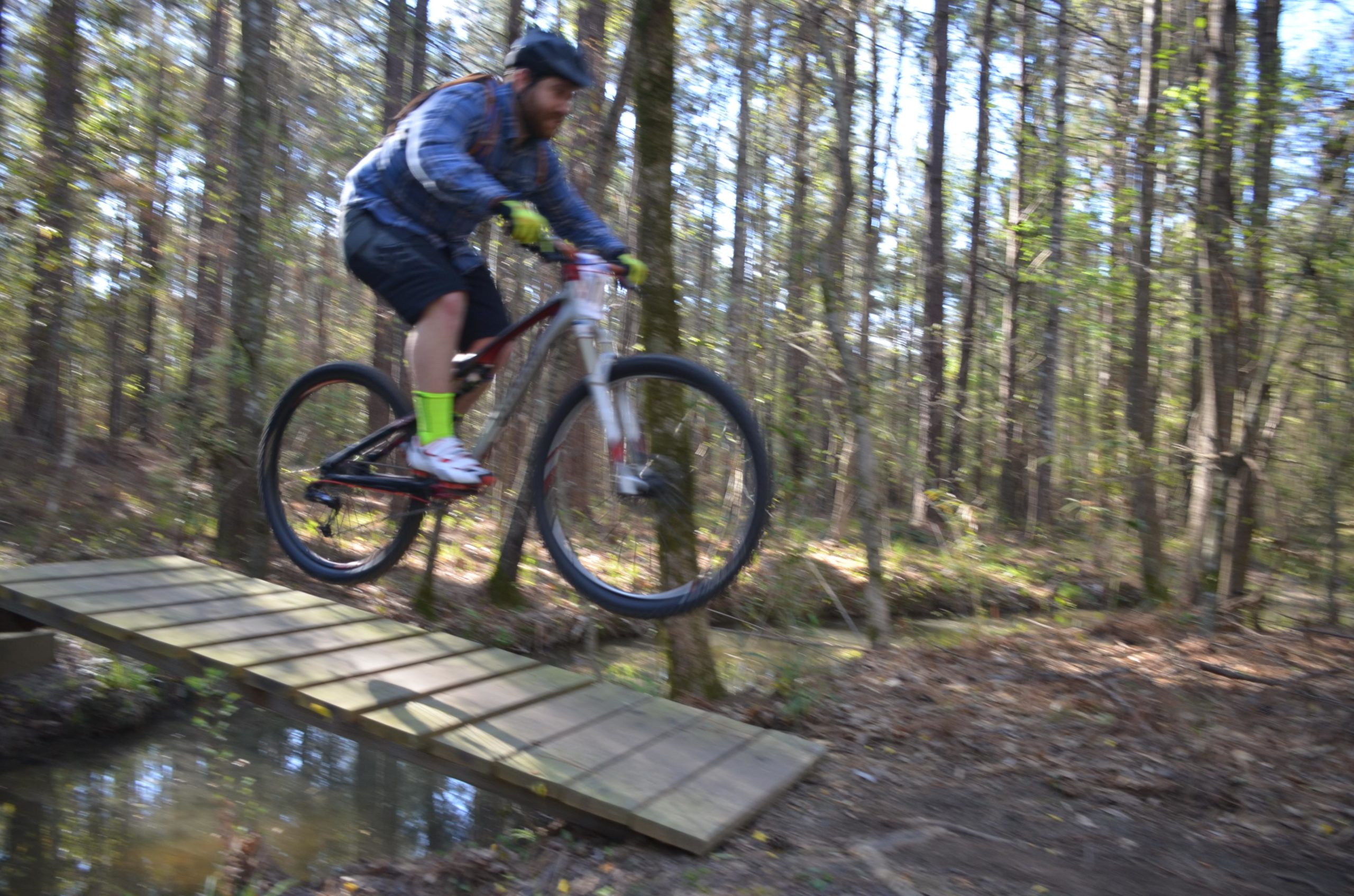 A mountain biker in mid-air, jumping over a wooden bridge in a forested area. The cyclist is wearing a helmet, a blue jacket, and bright green socks, with trees and underbrush visible in the background. Mt. Zion Bike Trails mountain bike trail.