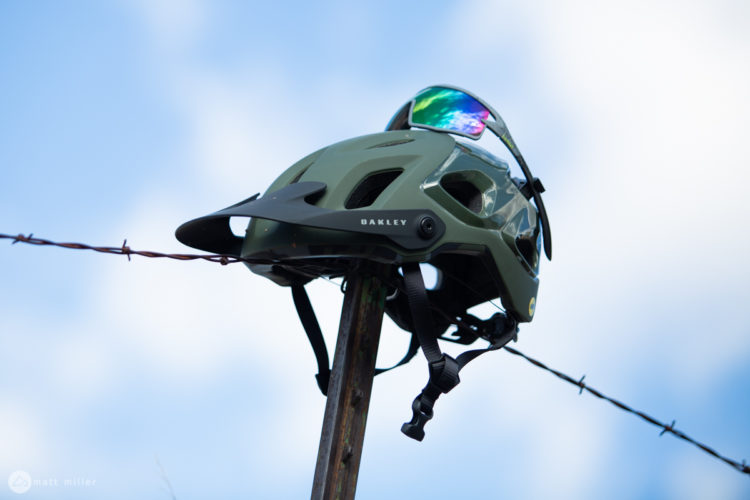 A green Oakley helmet perched on a wooden post, with barbed wire in the foreground and a blue sky in the background. The helmet features ventilation openings and a visor.
