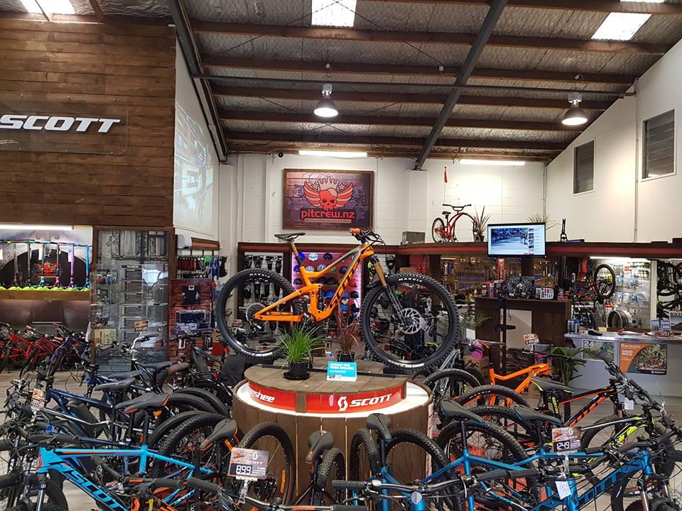 Interior view of a bike shop showcasing various bicycles, including prominently displayed models on a central podium. The shop features a mix of mountain bikes and accessories, with a wooden wall branding for "Scott" and a visually striking mural. Bright lighting highlights the vibrant colors of the bikes, and televisions display cycling content in the background.