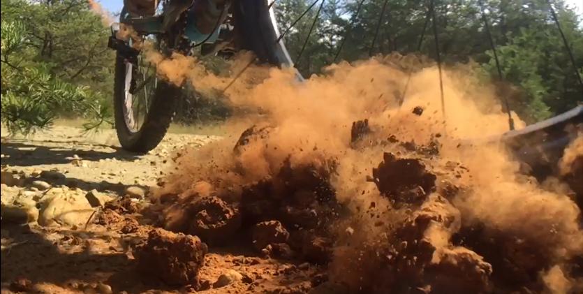 A close-up view of a mountain bike tire as it rides over a dirt trail, kicking up clouds of dust and loose soil in the air. The background features trees, indicating a natural outdoor environment. Glouster County college. mountain bike trail.