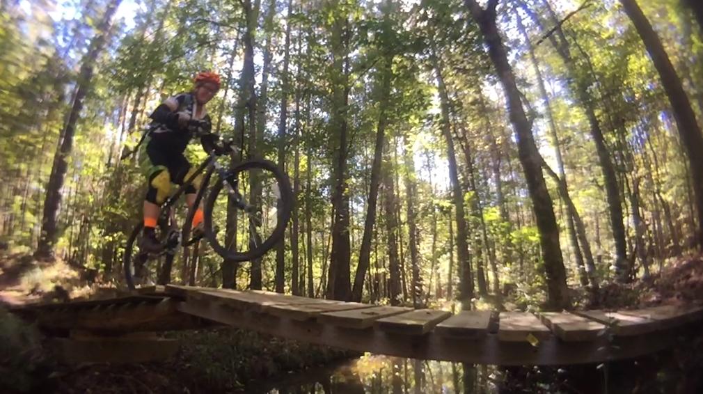 A mountain biker in mid-jump crosses a wooden bridge over a small stream, surrounded by lush greenery and tall trees in a vibrant forest setting. The biker wears a helmet, gloves, and brightly colored shorts, showcasing an adventurous spirit in an outdoor environment. Mt. Zion Bike Trails mountain bike trail.