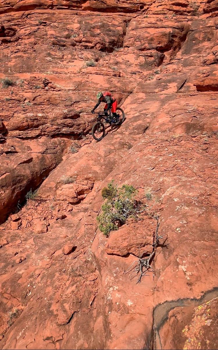 A mountain biker navigating a rocky trail on a steep red rock surface, with sparse vegetation in the background. The biker is wearing a helmet and bright red gear, showcasing an adventurous spirit in an outdoor setting. Hiline mountain bike trail.