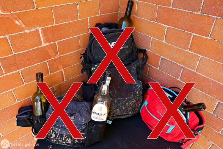 Three backpacks are positioned against a brick wall, each holding bottles that are crossed out with red "X" marks, indicating they are not allowed. The backpacks vary in color and style, one is black and heavily soiled, while another is red with turquoise accents.