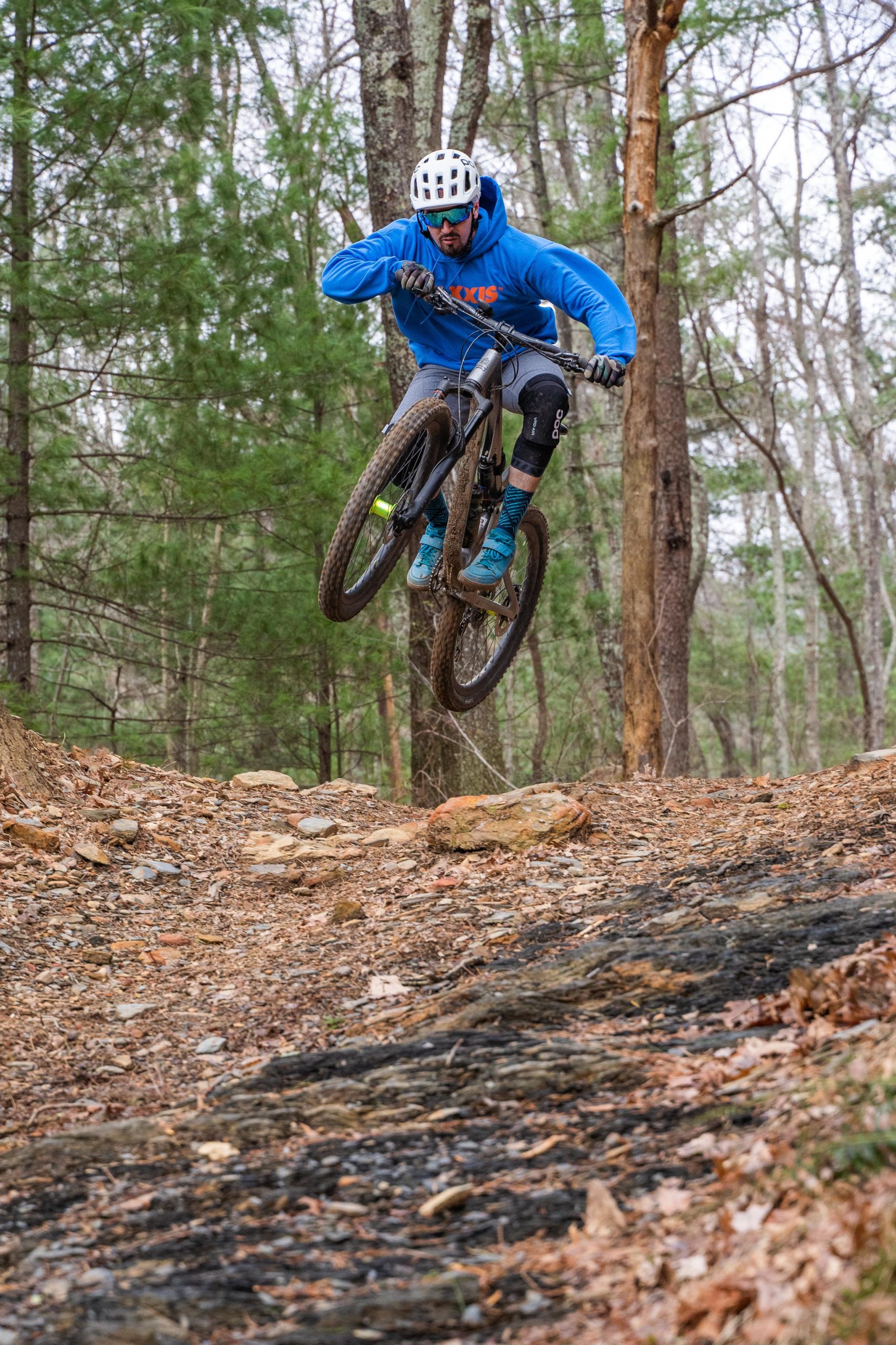 A mountain biker performing a jump on a dirt trail surrounded by trees, showcasing a dynamic action pose. The rider is wearing a blue hoodie, black shorts, knee pads, and a helmet, with a focused expression as they navigate the rocky terrain. Windy Gap Trail mountain bike trail.