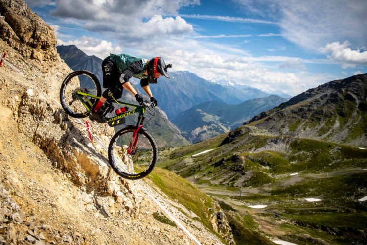 A mountain biker navigates a rocky slope in a mountainous landscape, showcasing skill and balance. The backdrop features rolling hills and distant peaks under a blue sky with scattered clouds.