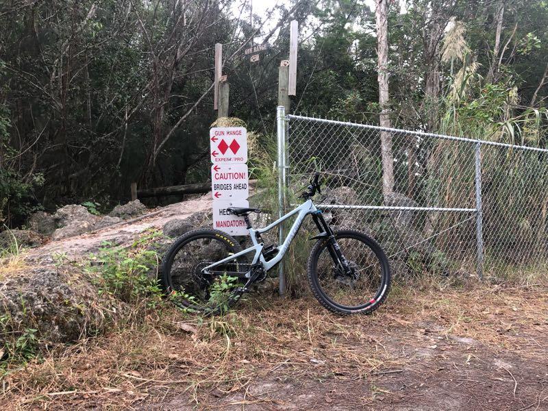 A mountain bike leaning against a fence near a sign that warns of a nearby gun range and advises caution regarding bridges ahead. The scene is surrounded by dense greenery and a dirt path. Markham Park mountain bike trail.