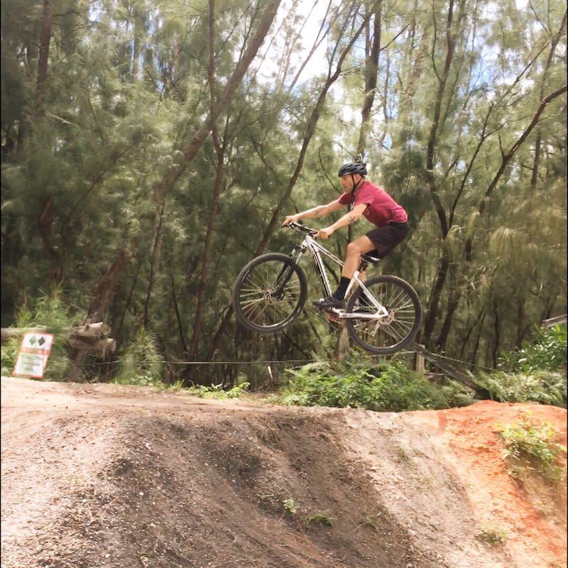 A person in a red shirt and black shorts is riding a mountain bike and jumping off a dirt ramp in a wooded area, with tall trees in the background. Oleta River State Park mountain bike trail.