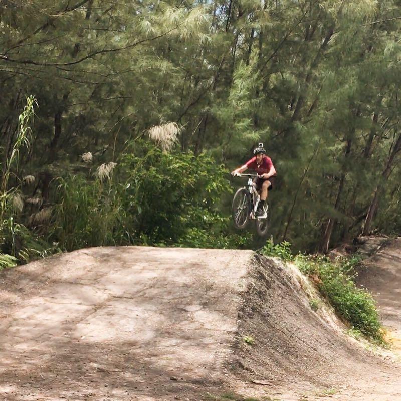 A cyclist mid-air performing a jump on a bike trail surrounded by lush greenery and trees. The rider is wearing a helmet and a red shirt, showcasing an adventurous moment in mountain biking. Oleta River State Park mountain bike trail.