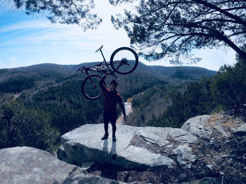 A person standing on a rocky outcrop, holding a bicycle above their head, with a scenic view of rolling hills and a river in the background. The individual is wearing a red and black checkered shirt and dark pants, surrounded by pine trees under a clear blue sky. Syllamo Trails mountain bike trail.