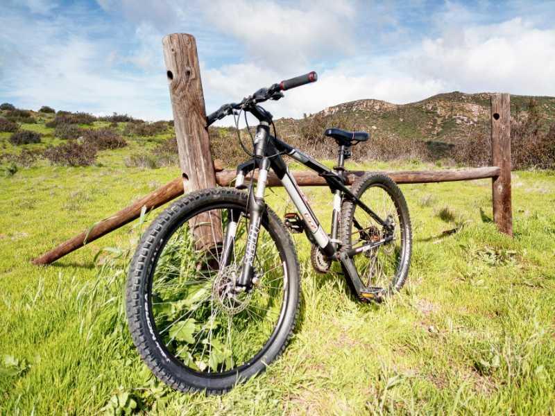 A mountain bike leaning against a wooden fence in a grassy field, with rolling hills and a partly cloudy sky in the background. Mission Trails mountain bike trail.