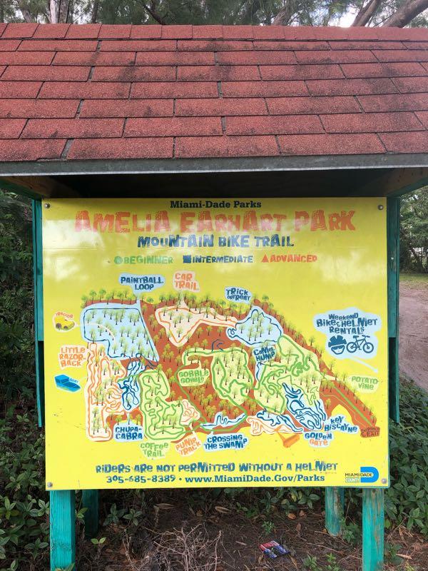 Sign at Amelia Earhart Park displaying the mountain bike trail map, highlighting different trail difficulty levels: beginner, intermediate, and advanced. The map outlines various trails such as "Paintball Loop," "Cap Trail," and "Chupacabra," with notes on safety and bike rentals available on weekends. Bright colors and clear markings indicate different paths and features of the park. Amelia Earhart Park mountain bike trail.