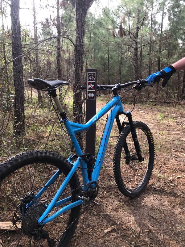 A blue mountain bike parked near a trail sign in a wooded area, featuring symbols for biking trails. The bike is positioned prominently, with a hand wearing a glove holding the handlebars. The background includes tall trees and a natural forest floor. Bogue Chitto State Park mountain bike trail.