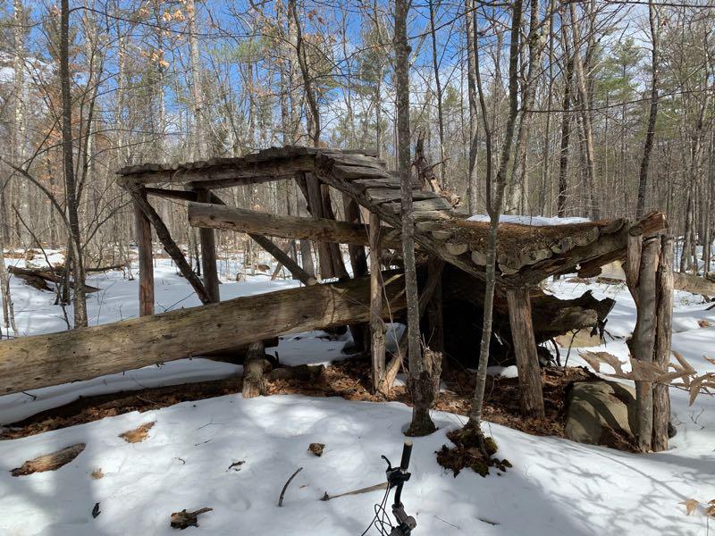 An old, weathered wooden structure partially collapsed in a snowy forest. The remains include a slanted roof covered with pine needles and moss, supported by various wooden beams. Surrounding the structure are sparse trees and a blanket of snow on the ground, with patches of exposed earth and fallen leaves visible. Bright blue skies peek through the tree branches above. Winant Park Hospital Trails mountain bike trail.