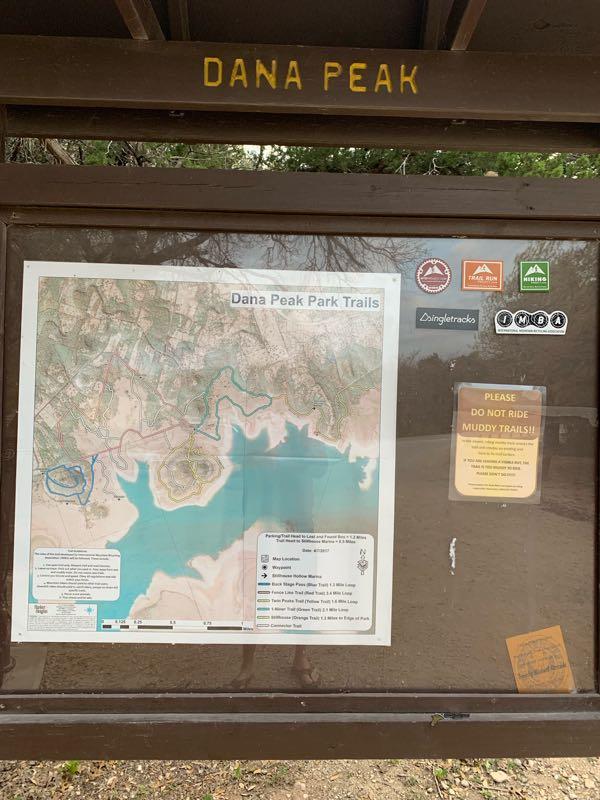 A wooden sign with the text "DANA PEAK" at the top, displaying a trail map of Dana Peak Park. The map includes various trails marked in blue and green, surrounding a body of water. There are several informational and warning signs regarding trail conditions and navigation, alongside various park logos. The area appears to be a natural outdoor setting. Dana Peak mountain bike trail.